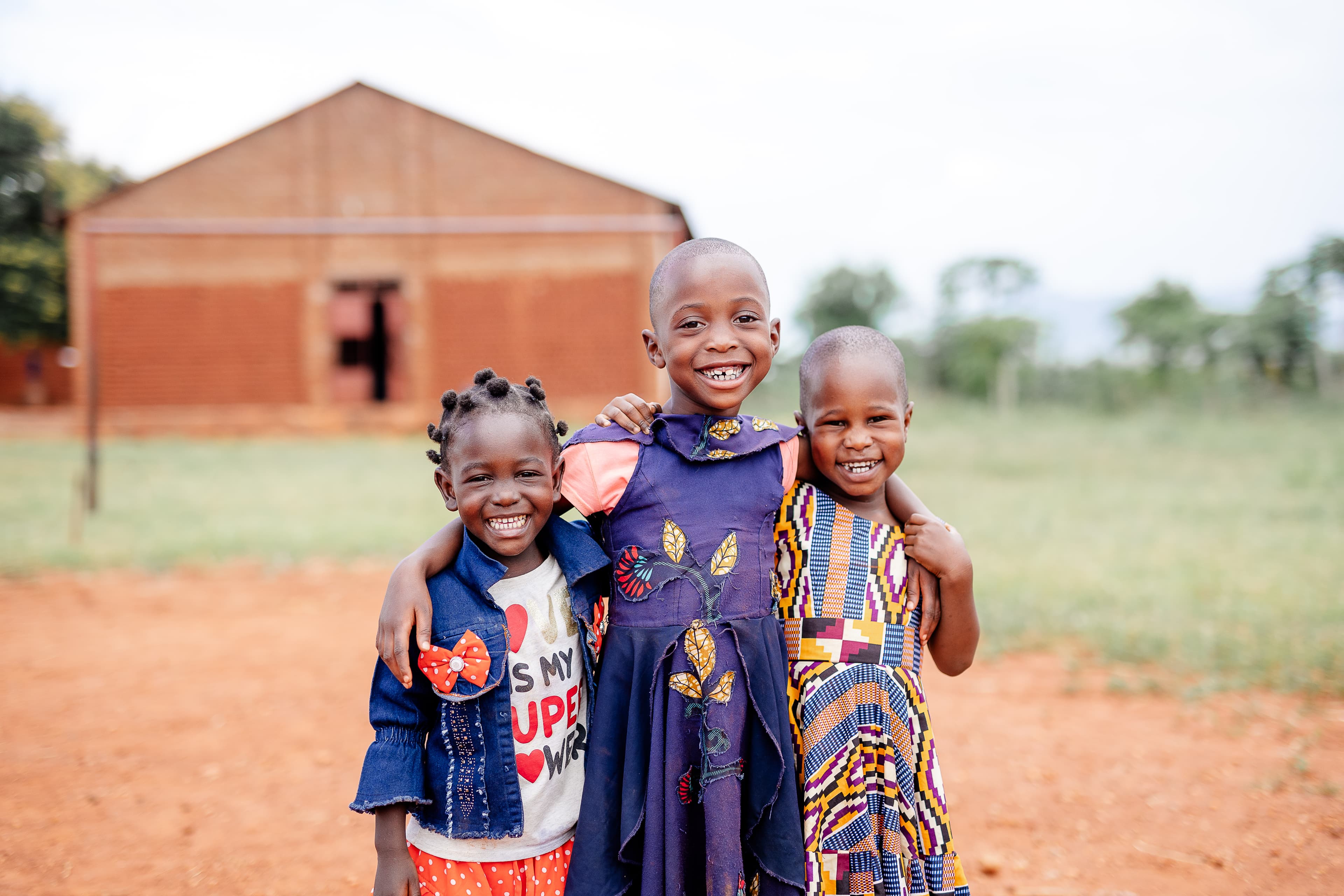 Three girls in colorful outfits stand outside in a red dirt field, smiling with their arms around each other.