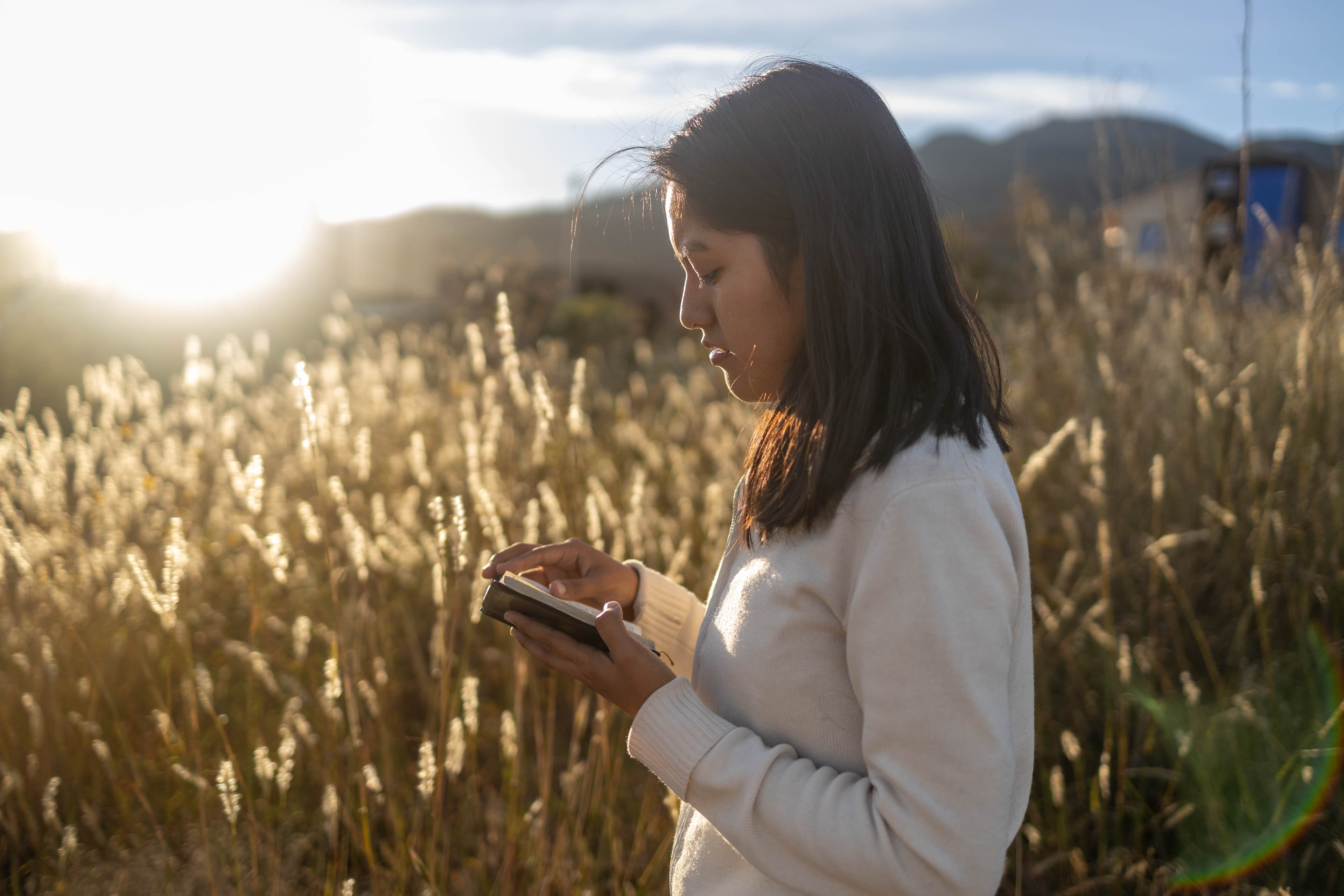 A woman in a cream sweatshirt, standing outside in a field, reading the Bible.