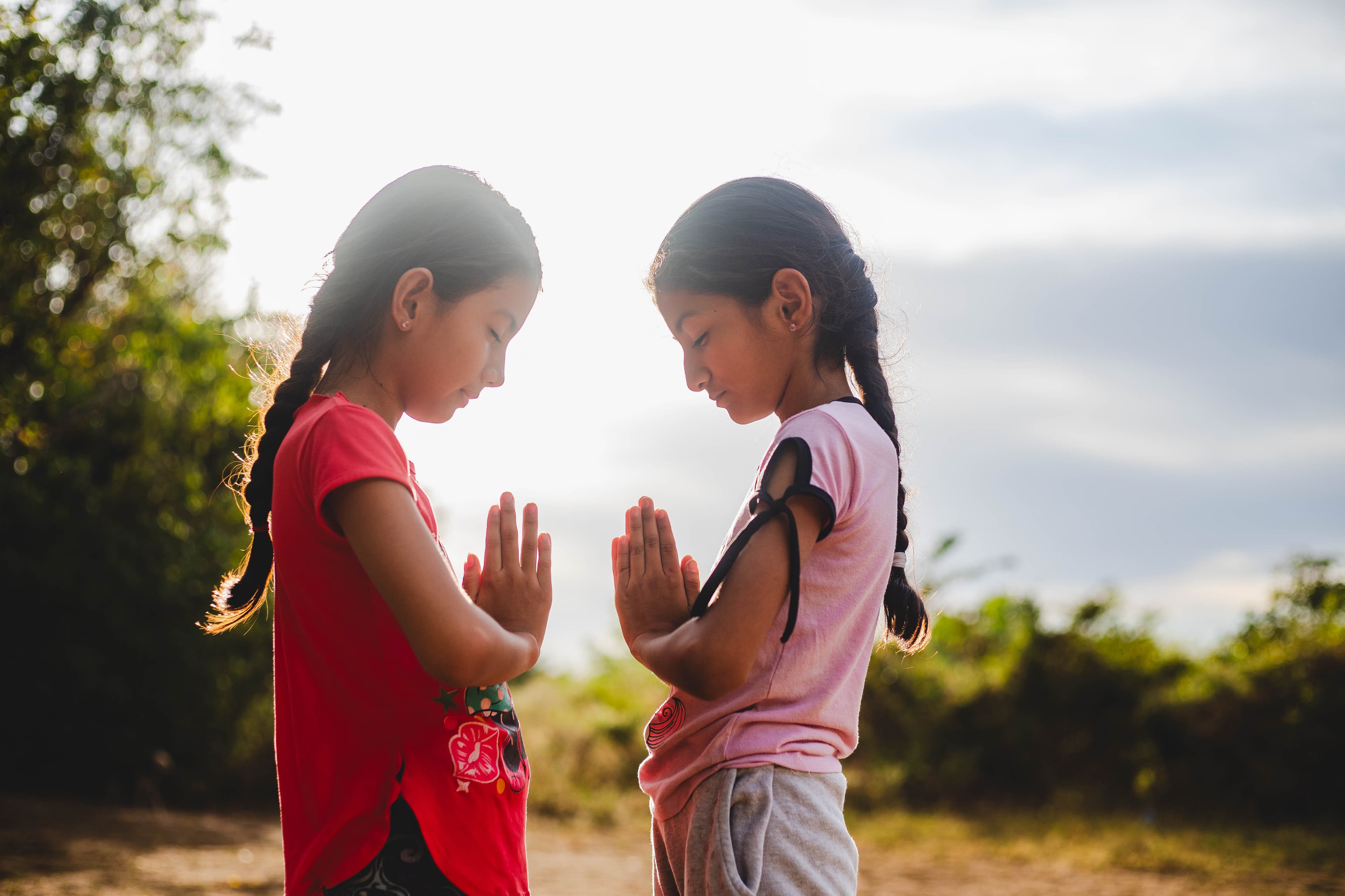 Two sisters are standing across from each other, their hands folded in prayer, with trees and vegetation in the background.
