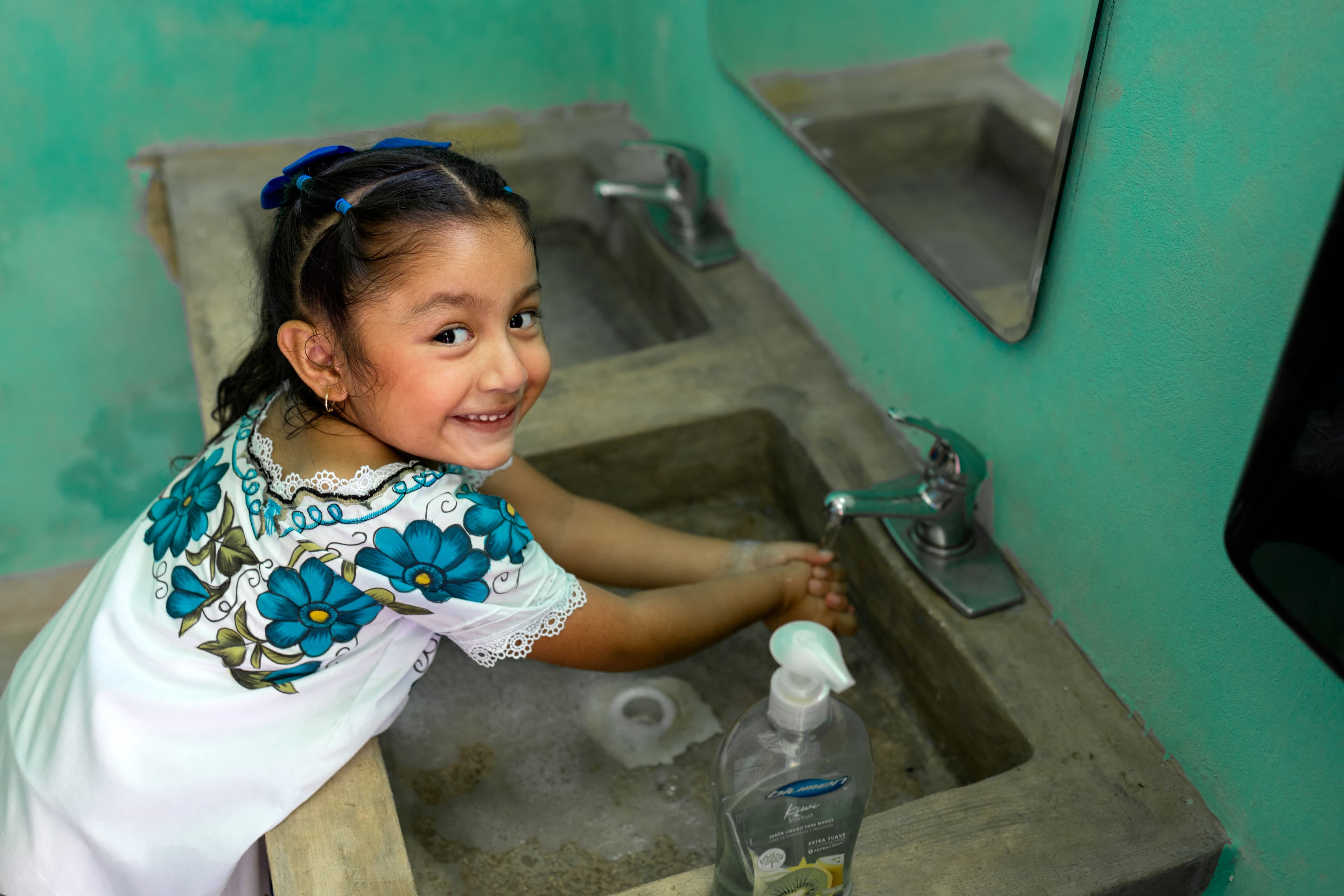 Young girl in a white and blue floral shirt smiles up at the camera while washing her hands.