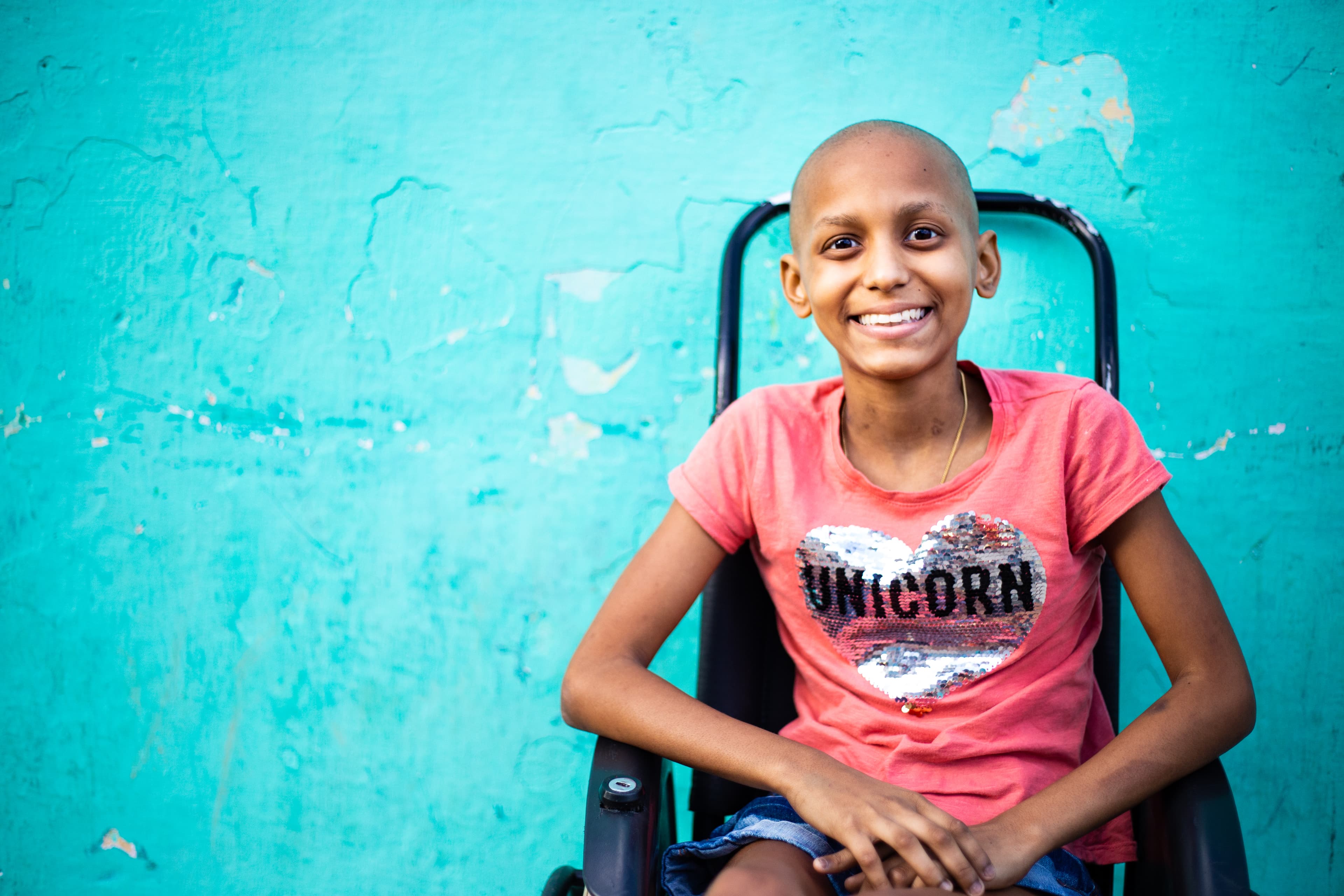 A pre-teen, female cancer patient smiles at the camera while sitting in her wheelchair. She is wearing a pink shirt.