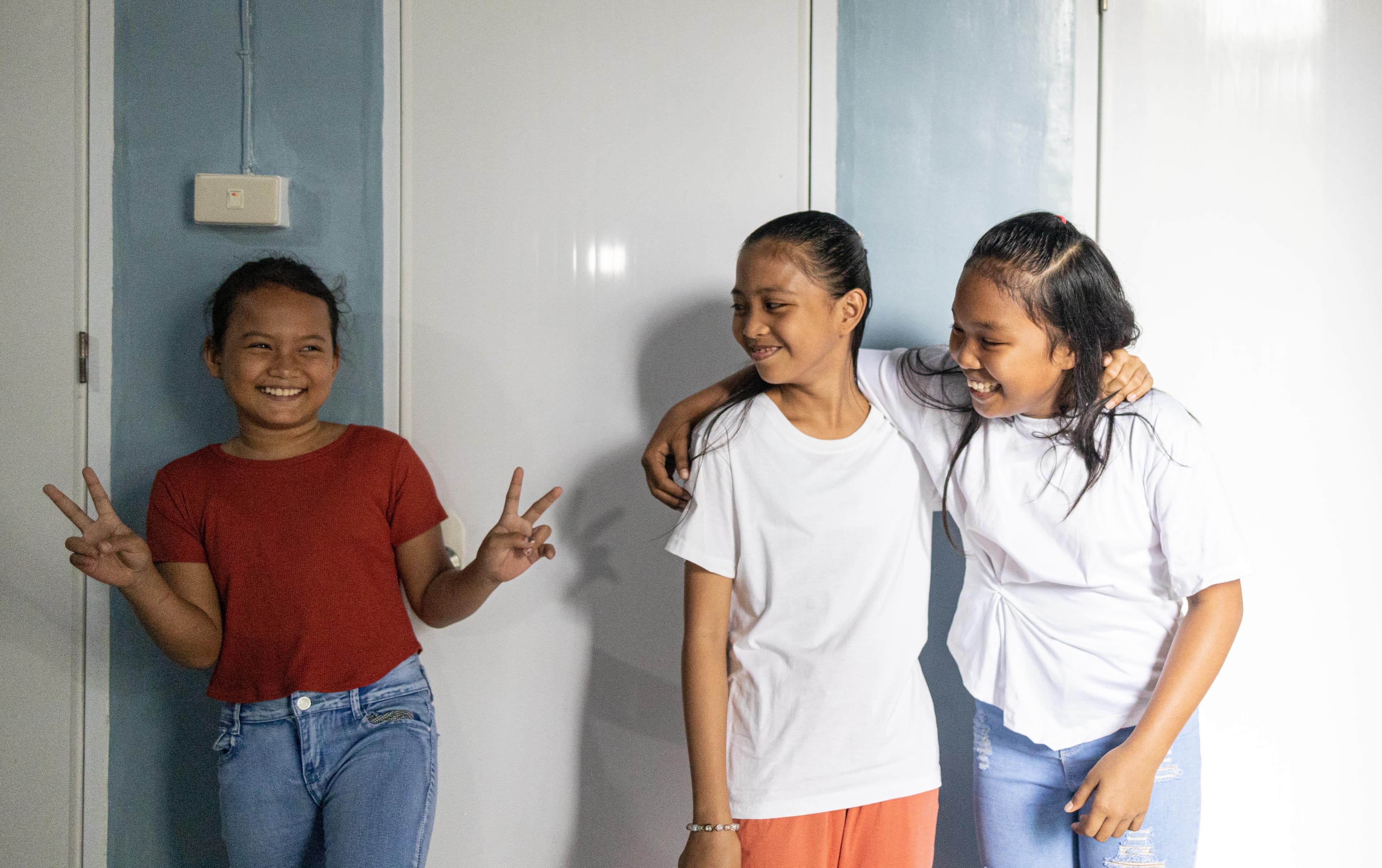 Two girls embrace while looking at another girl making peace signs with her hands.
