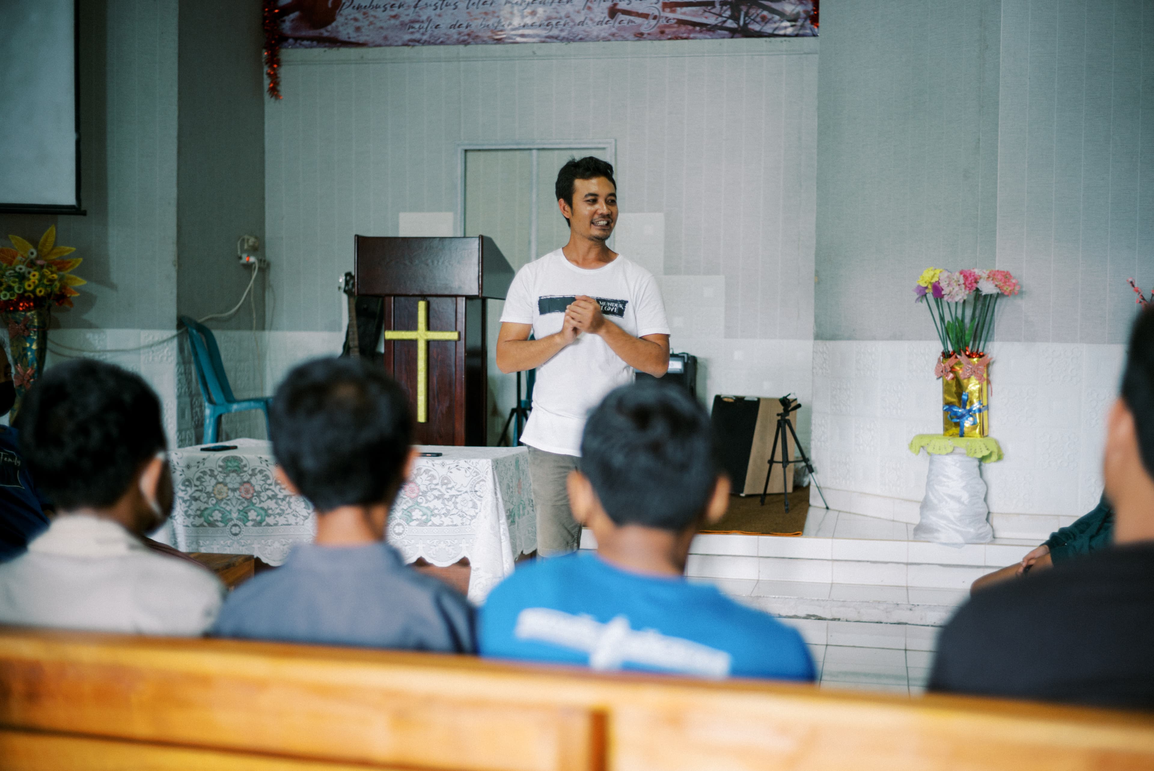 An adult man wearing a white shirt stands in front of a church while speaking.