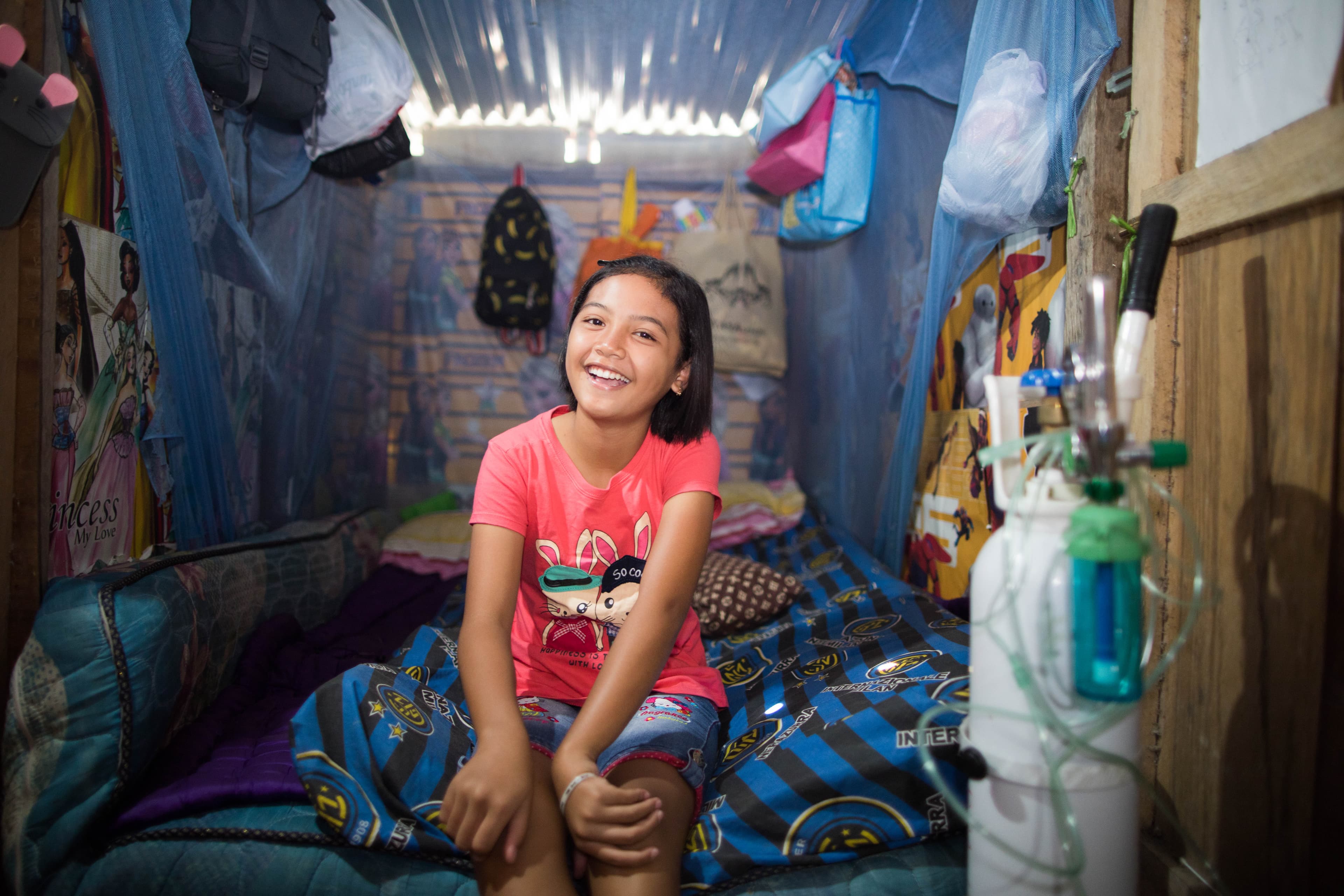 A young girl is sitting on the end of her bed smiling next to her oxygen tank.