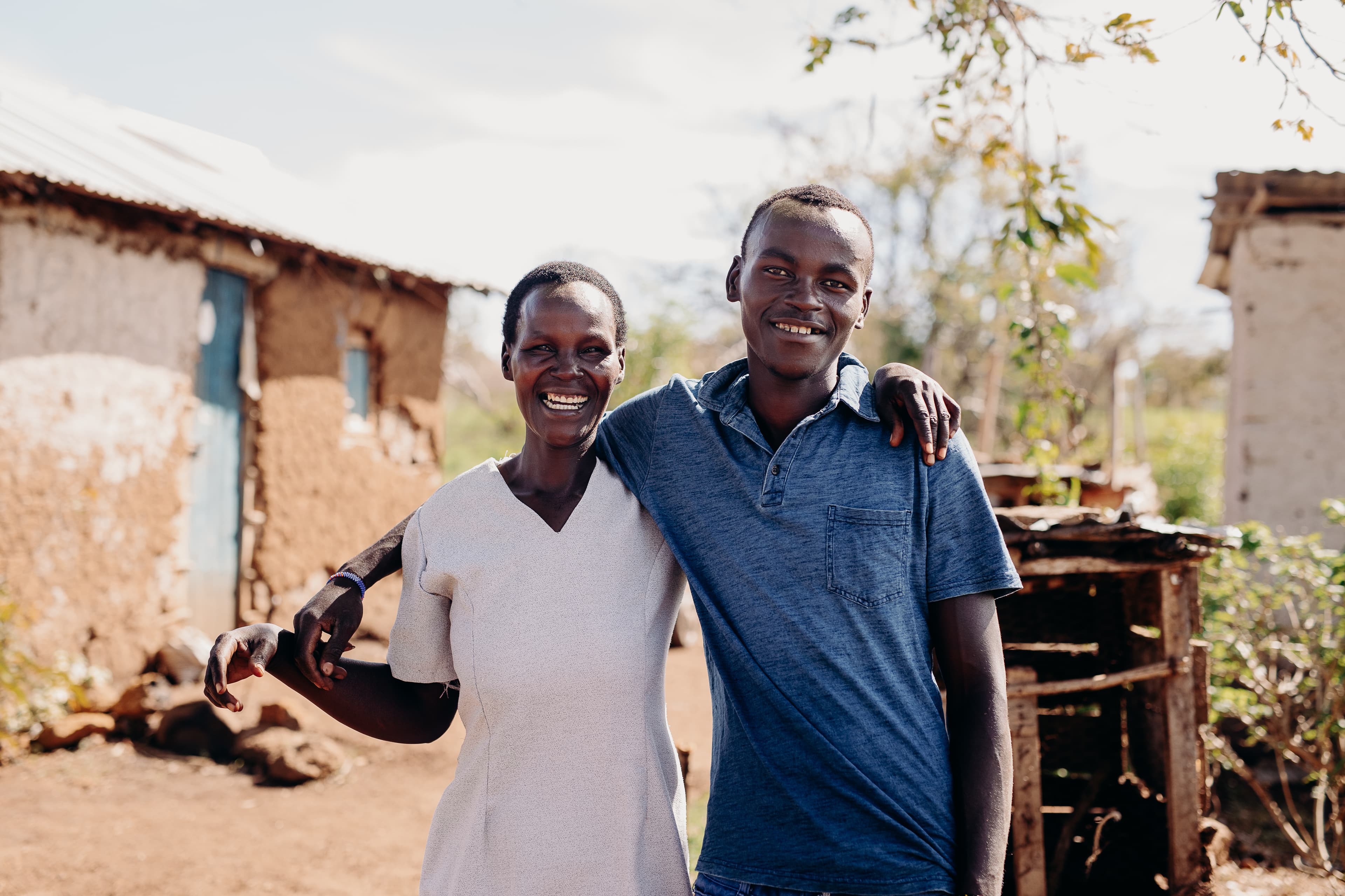 A Kenyan teen and his mother link arms while smiling for the camera.