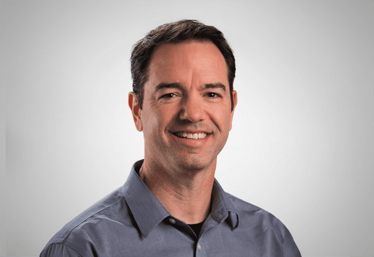 A man in a grey button up shirt smiles, with a grey background behind him.
