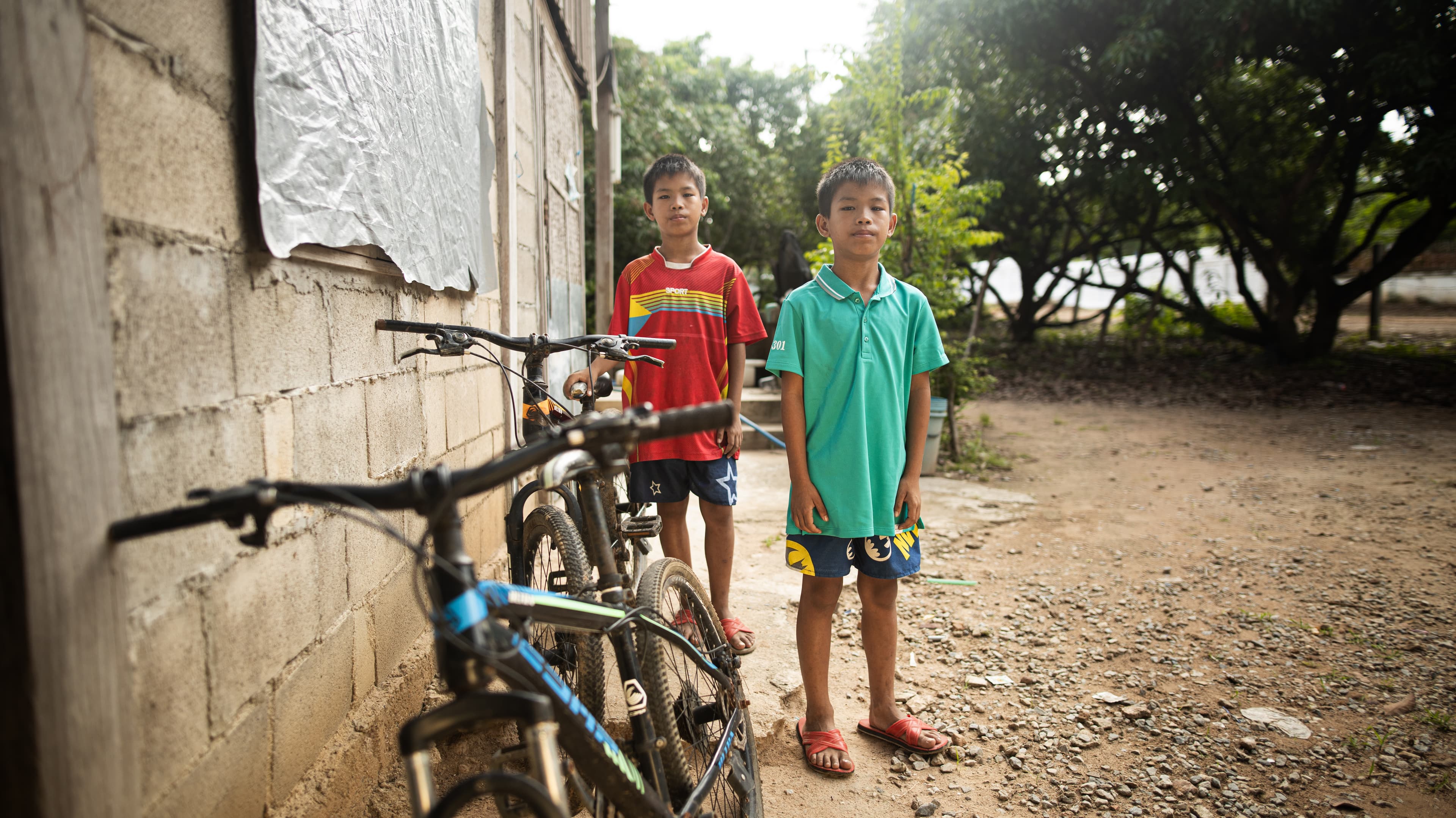 Two Thai boys stand outside their block home behind two bicycles.