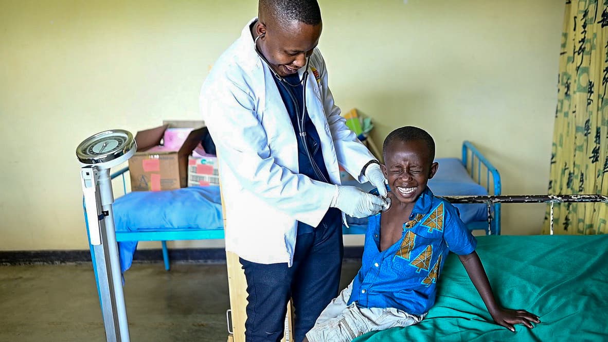 An African doctor wearing a white lab coat checks the heart of a young African boy with a stethoscope.