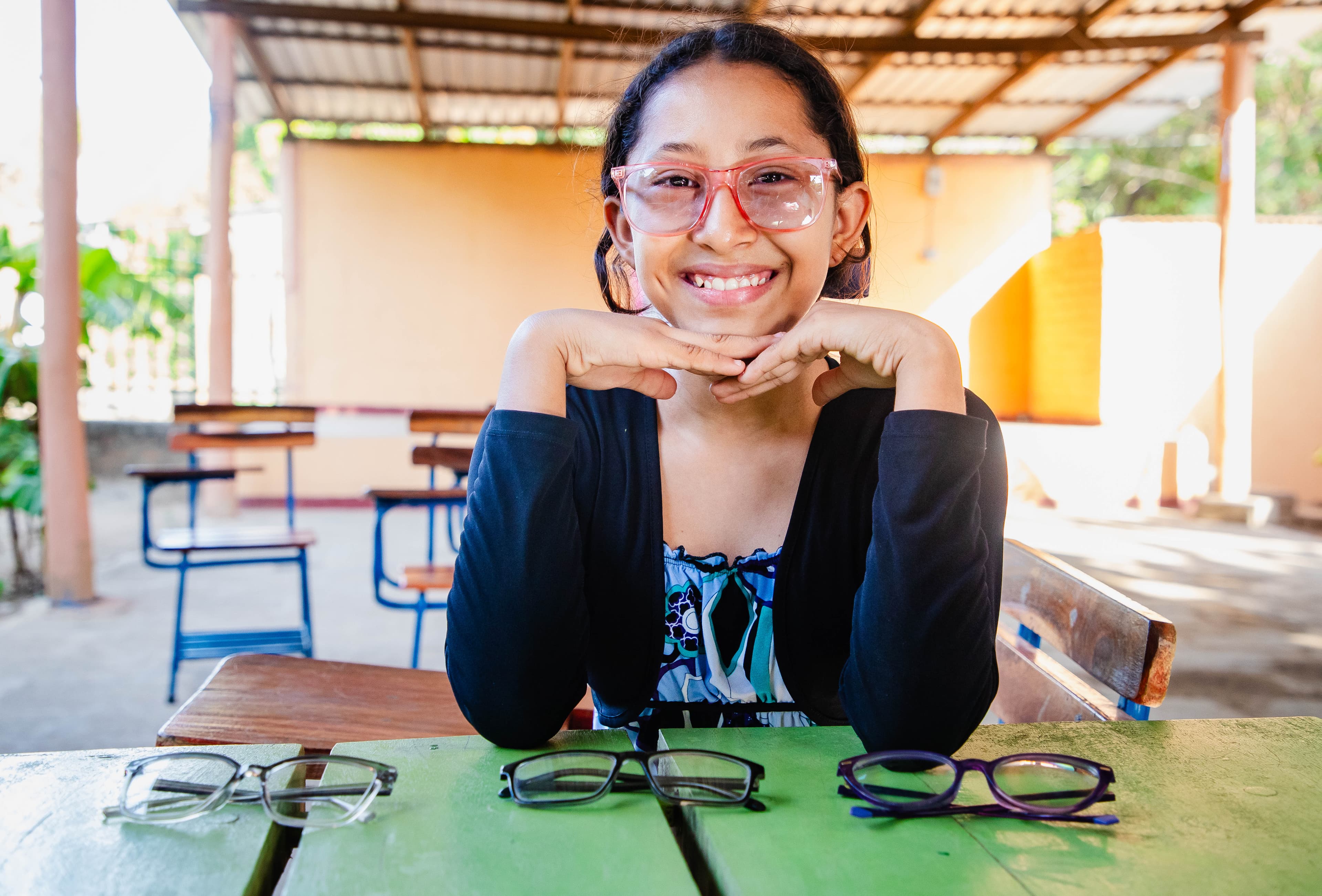 A young girl wearing glasses sits at a green table with her head resting on her hands.