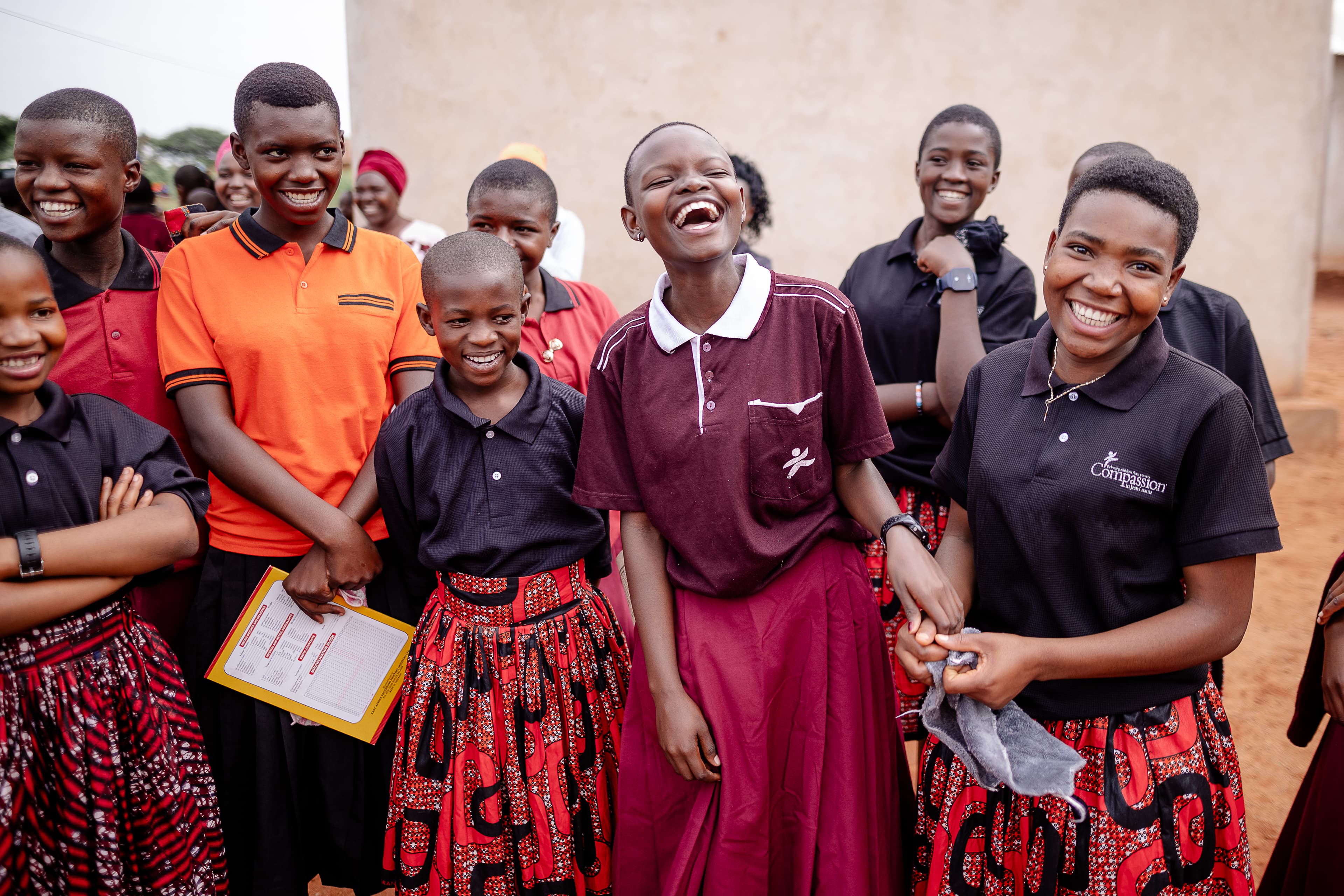 A group of children and adults gather together, smiling and laughing outside a concrete building. They are wearing polo shirts in colors of orange, blue and burgundy.