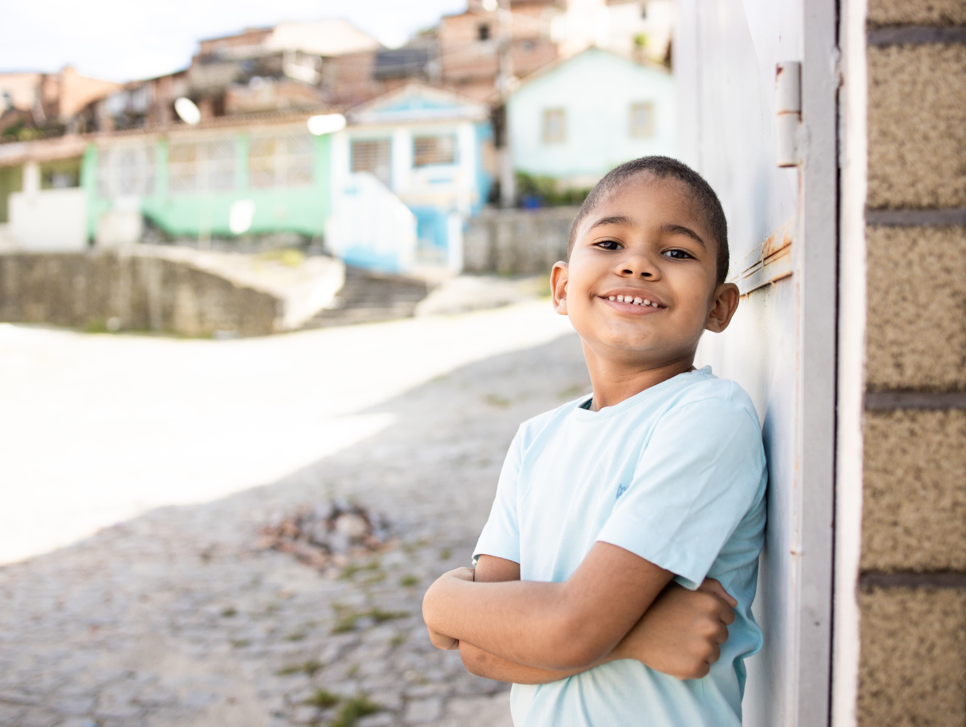 A young Brazilian boy leans against a wall with his arms crossed as he smiles for the camera.