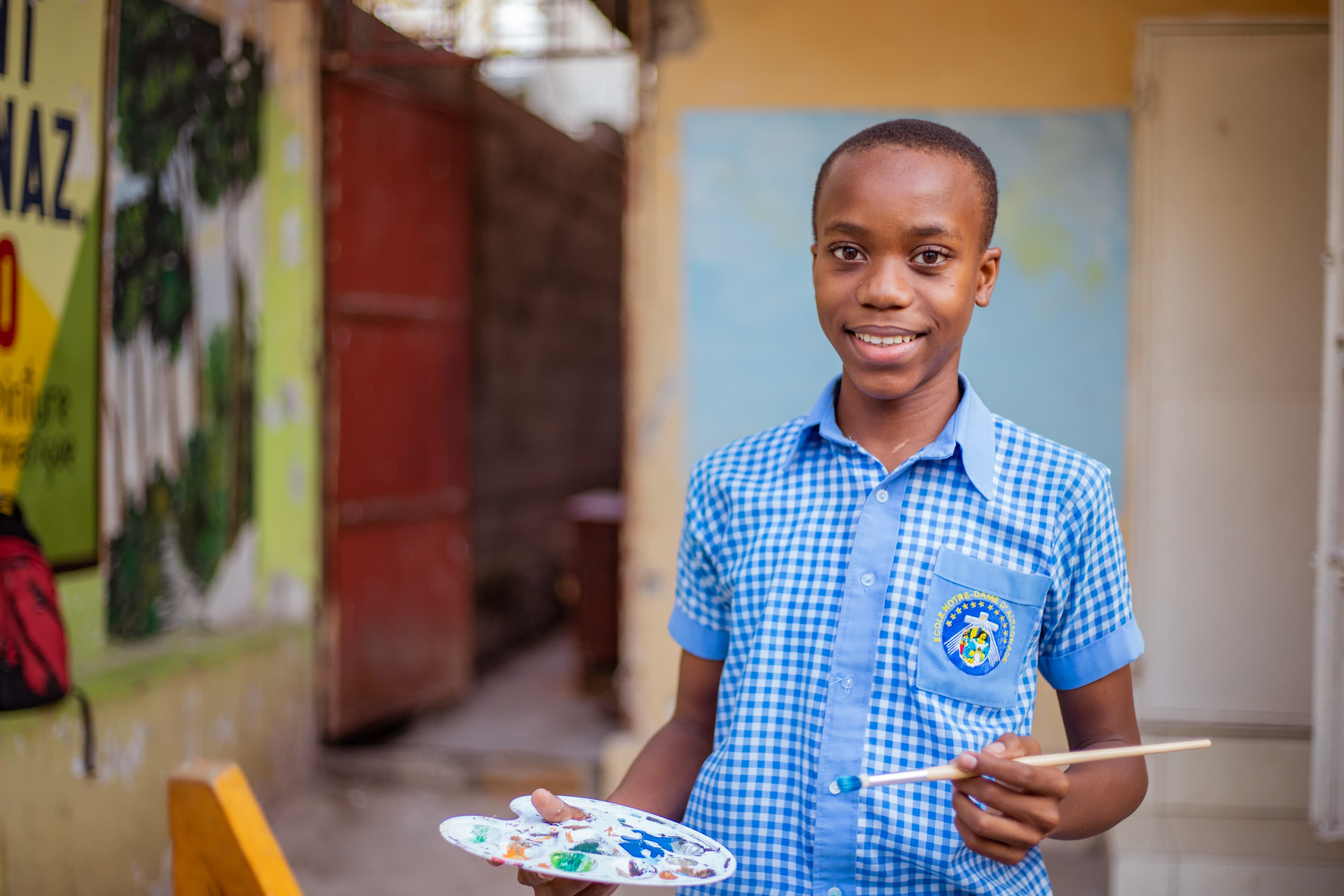A teenaged boy wearing a blue and white checkered shirt holds a paintbrush and palette.