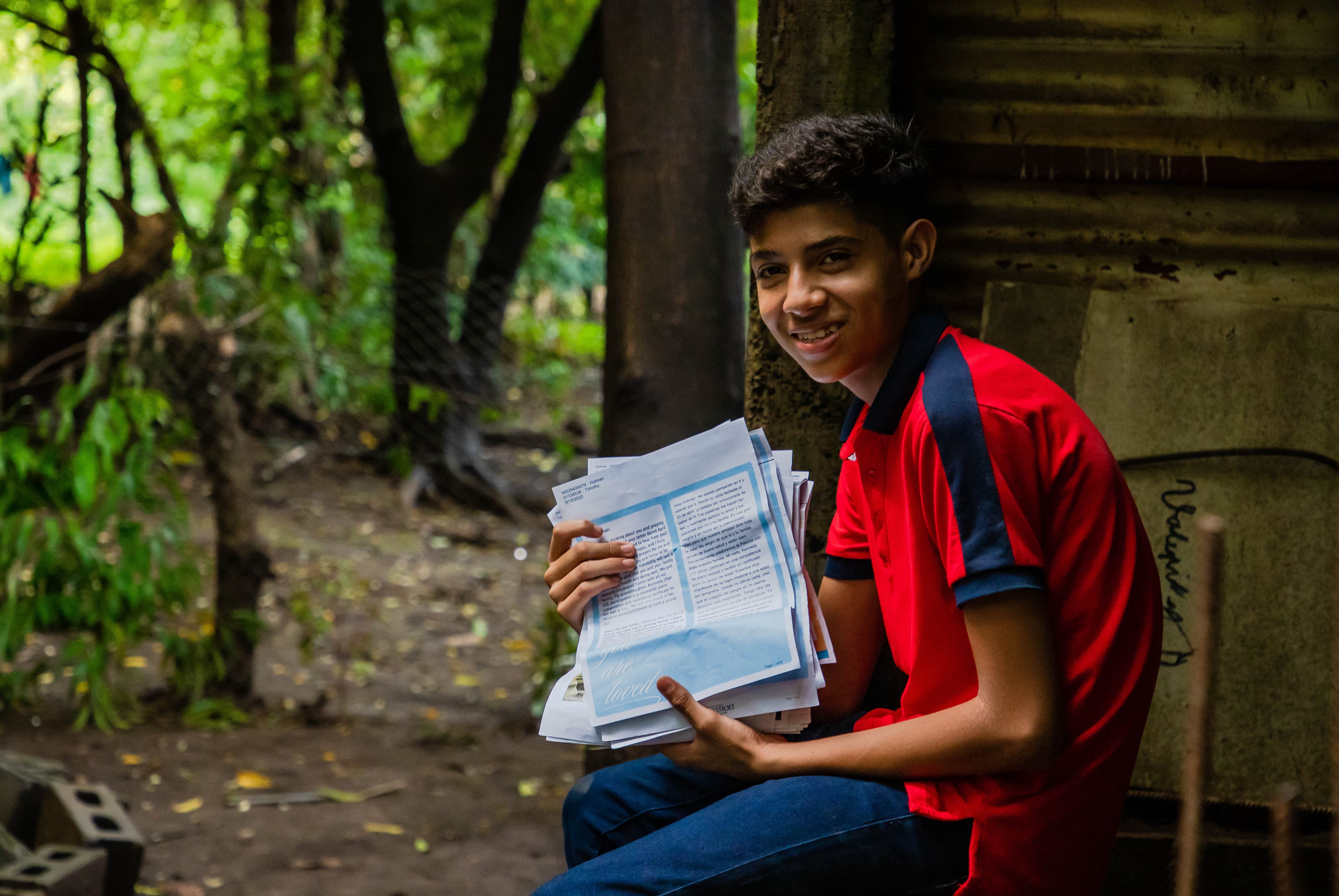 Boy in red and blue polo smiling at camera while holding stack of letters outdoors.