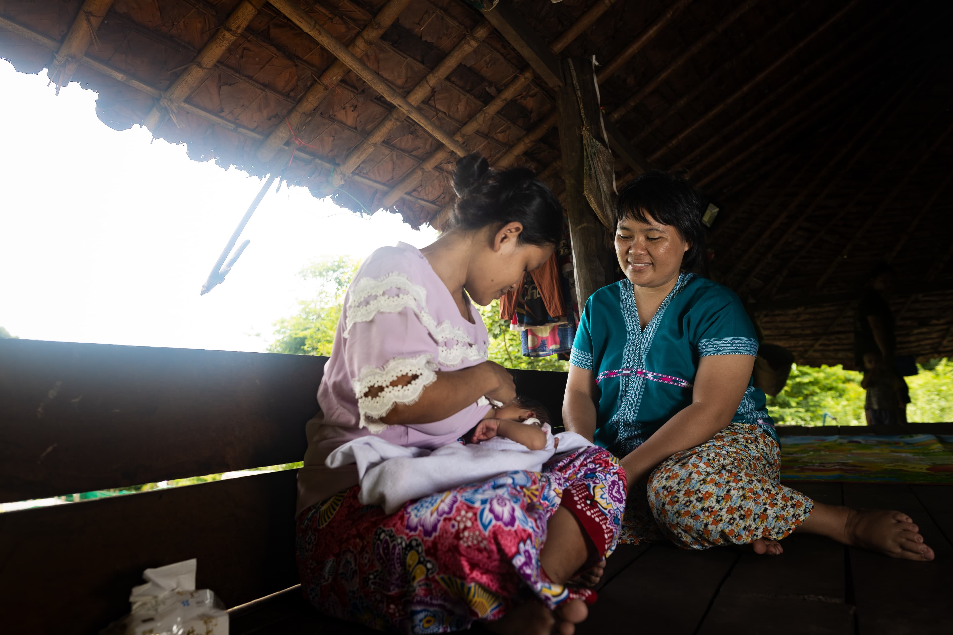 A mother sits on a bench holding her baby while a woman sits beside her in support.