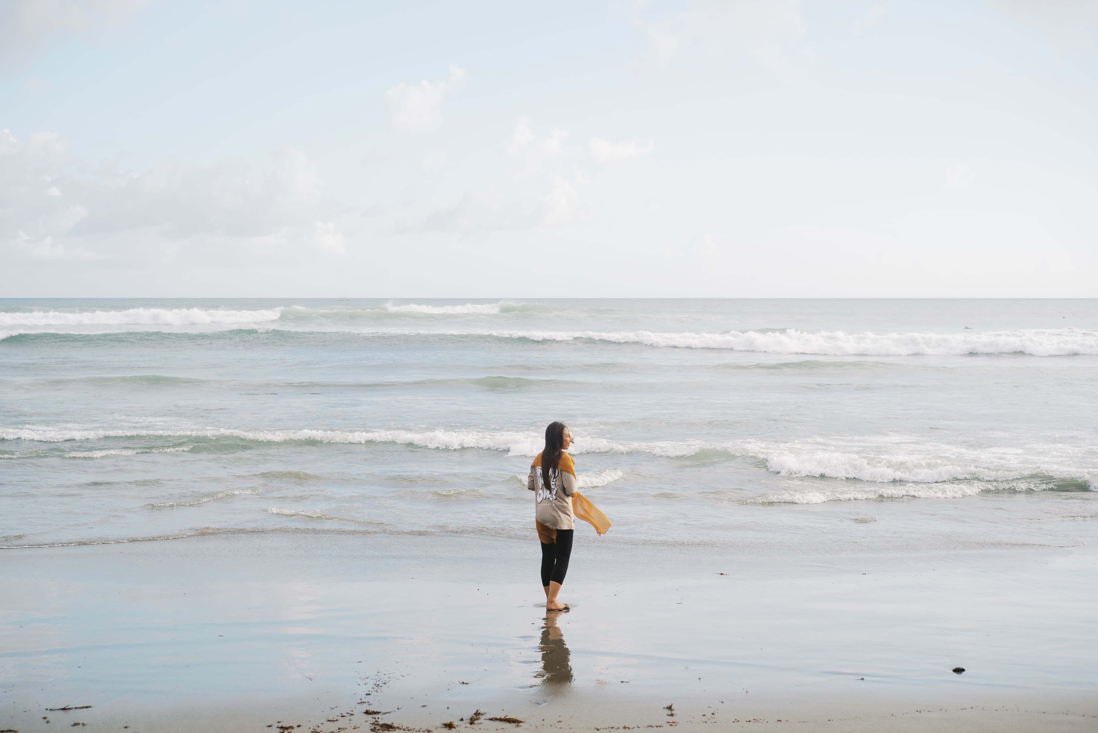 A teen Indonesian girl stands on a beach holding a gold scarf with waves and sky in the background.