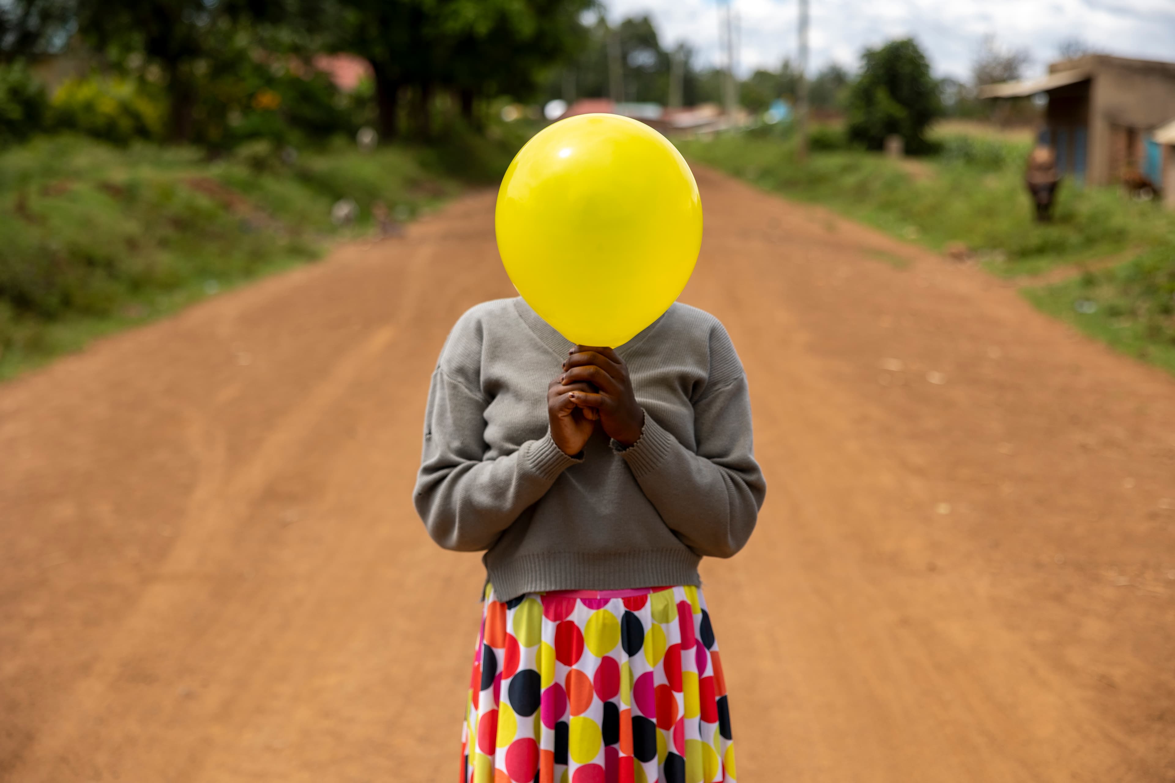 A young girl holds a yellow balloon in front of her face while standing on a dirt road.