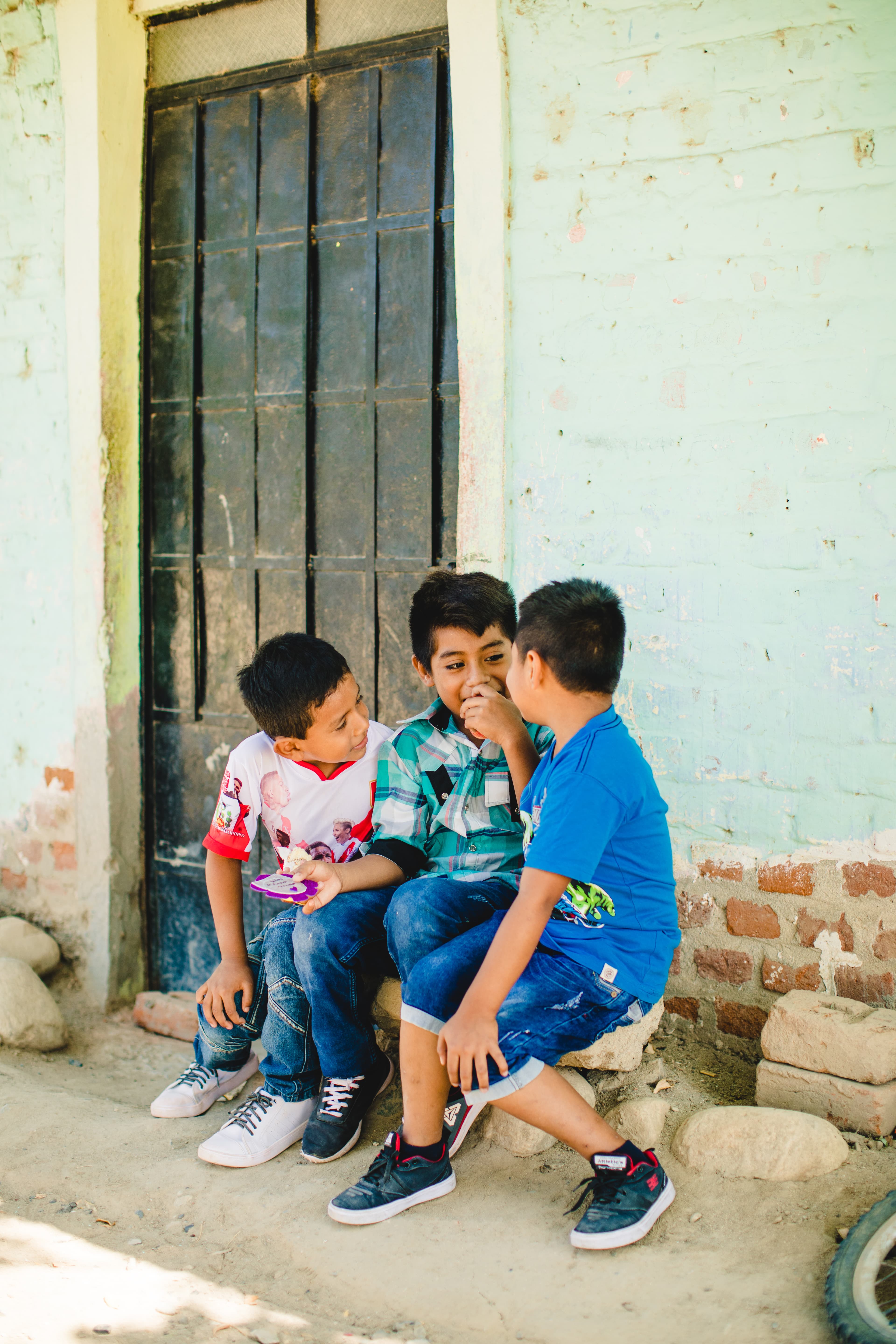 Three boys in red and white, green and blue shirts sit in front of a pale green building with a black door.