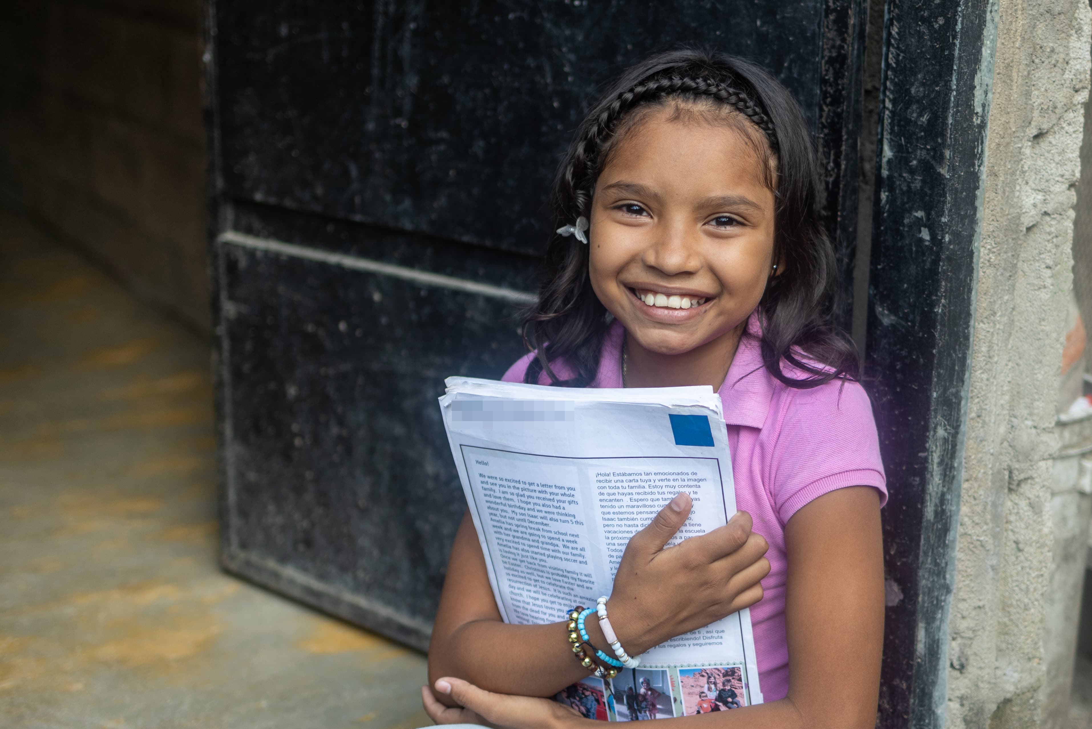 A young girl holds letters while smiling and leaning against a building.