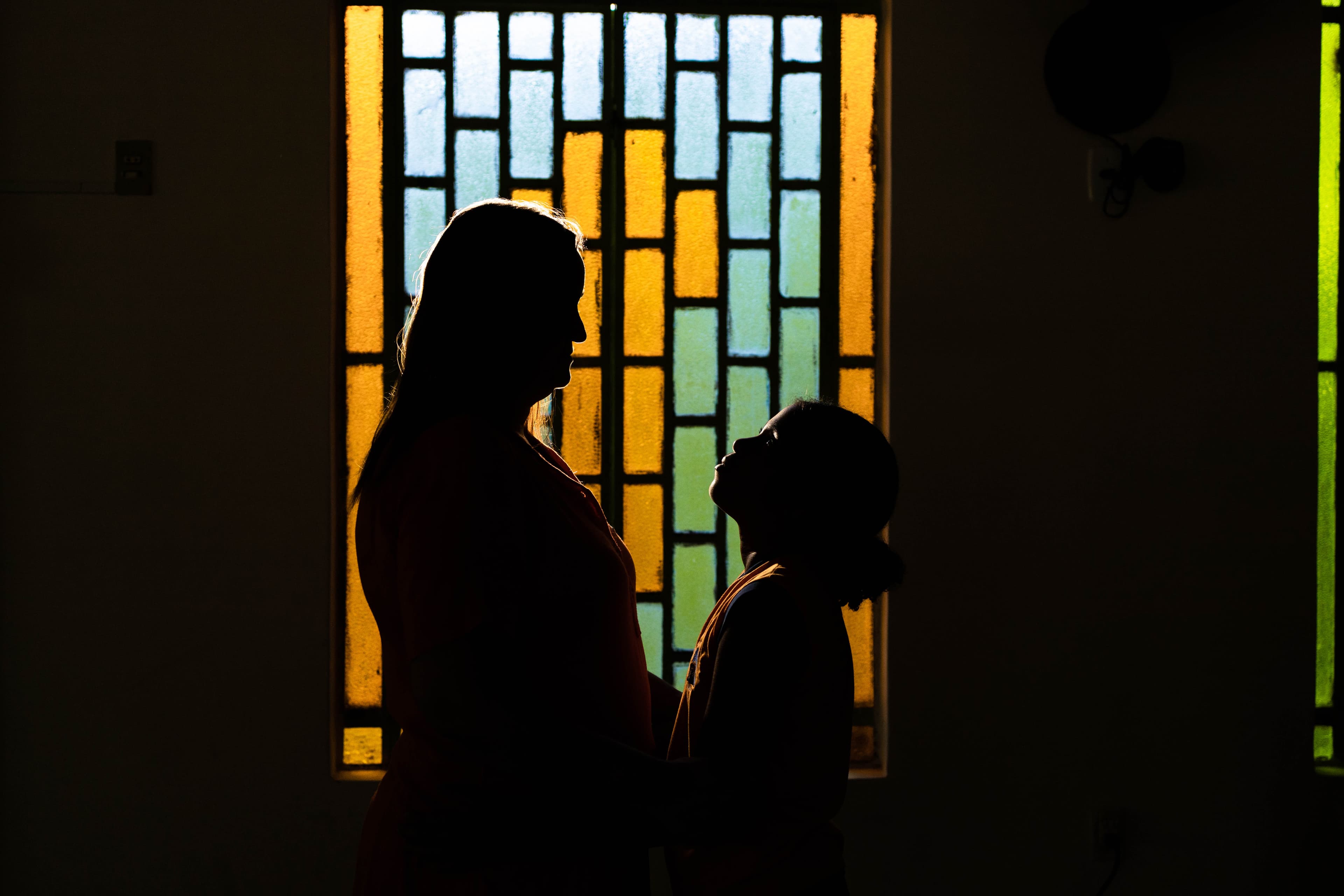 An older woman and a young girl stand in front of a stained glass window with their faces hidden.