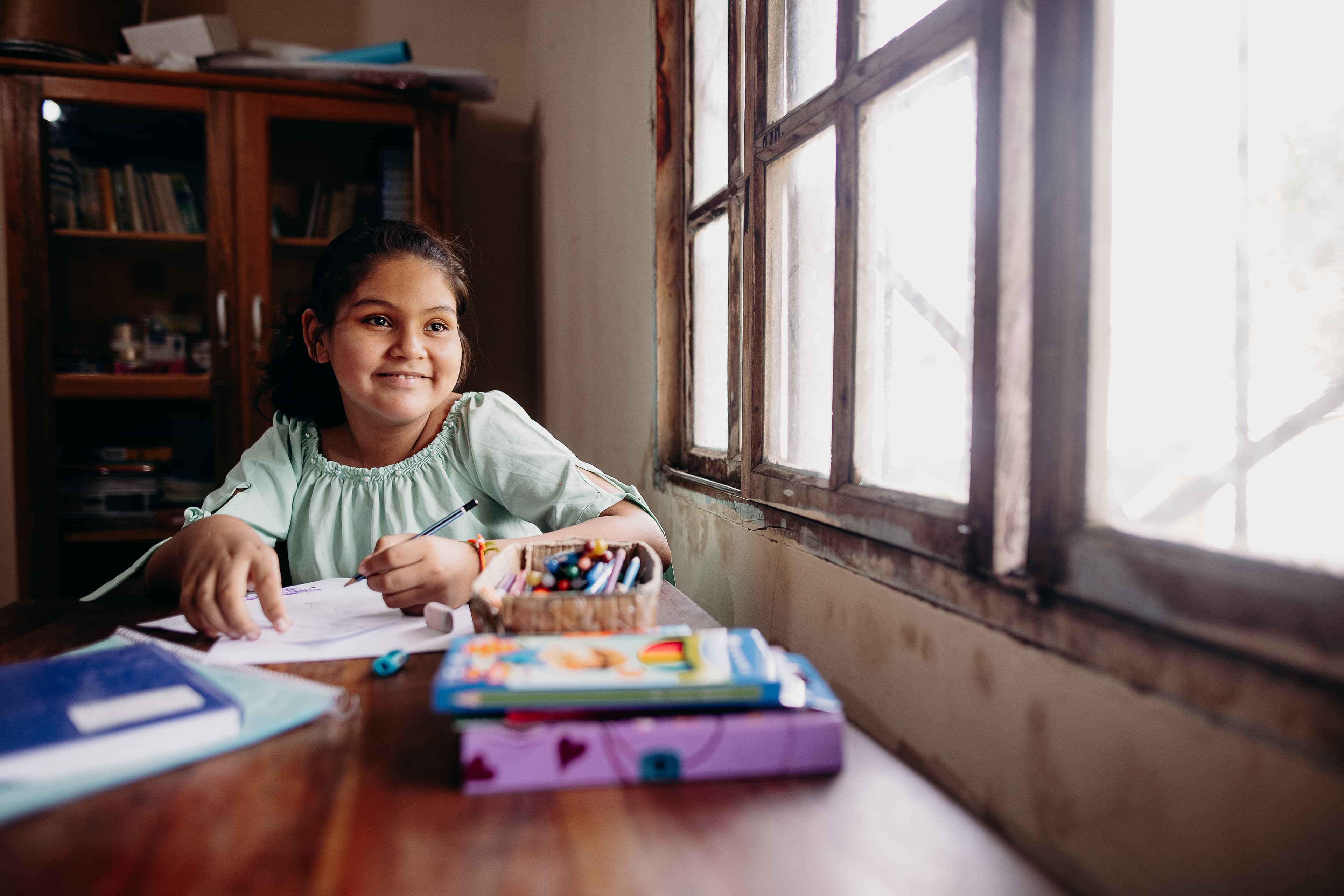 A young girl holds a pen and smiles while looking out a window.