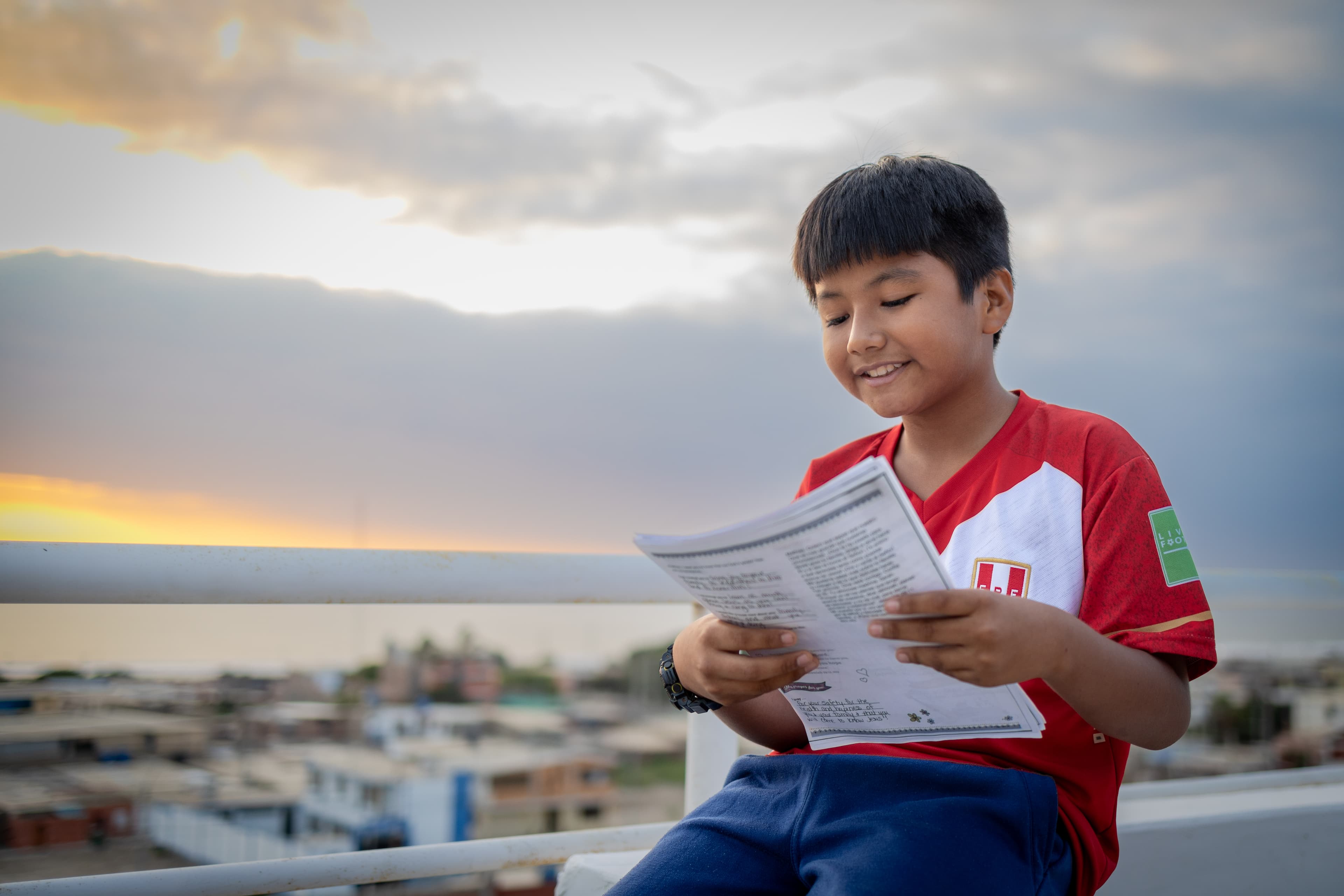 A boy wearing a red and white shirt is sitting outside reading a letter. He is smiling. There are clouds, sunset and a neighborhood behind him.