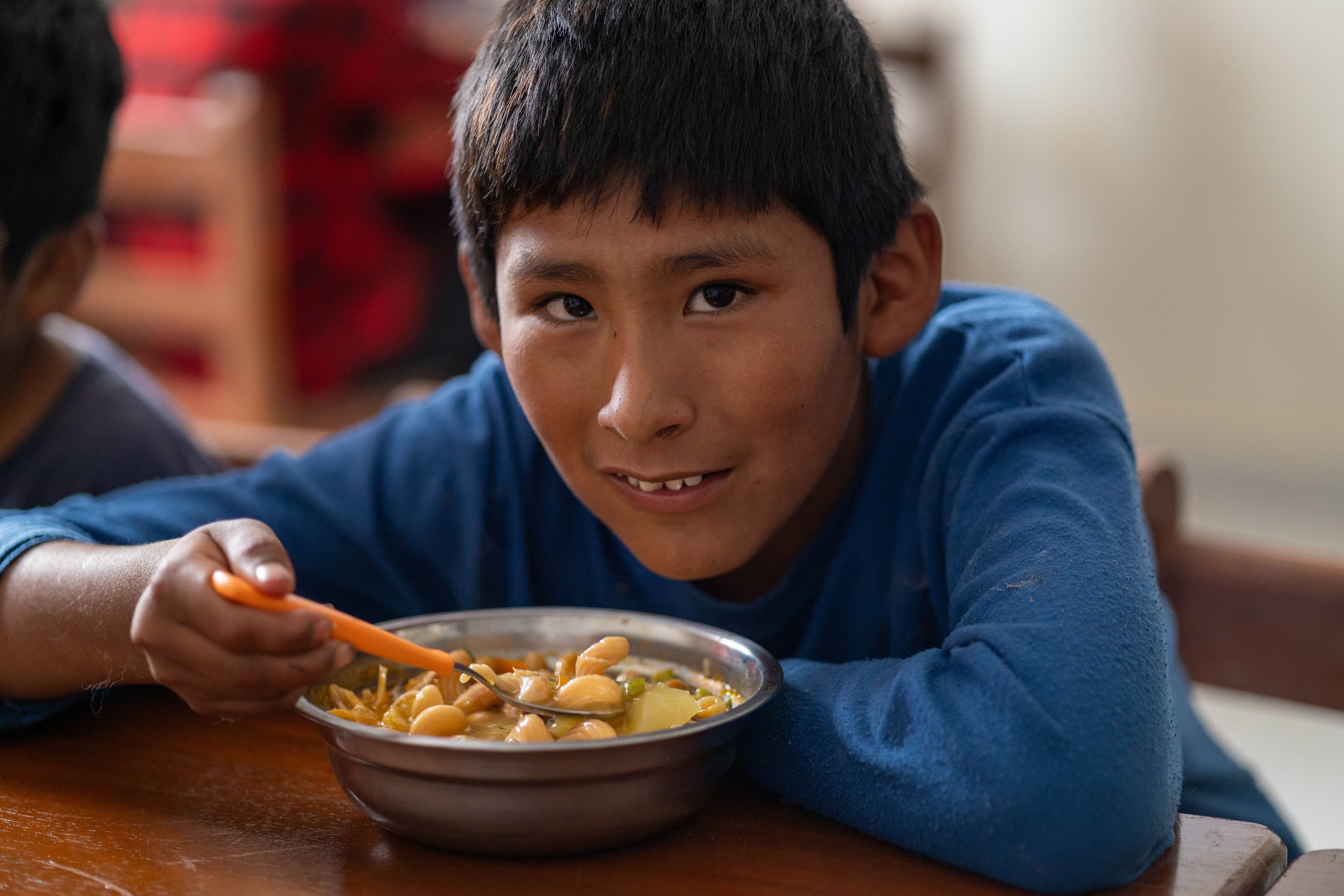 A boy in a blue shirt sits at a wooden table, eating soup from a metal bowl.