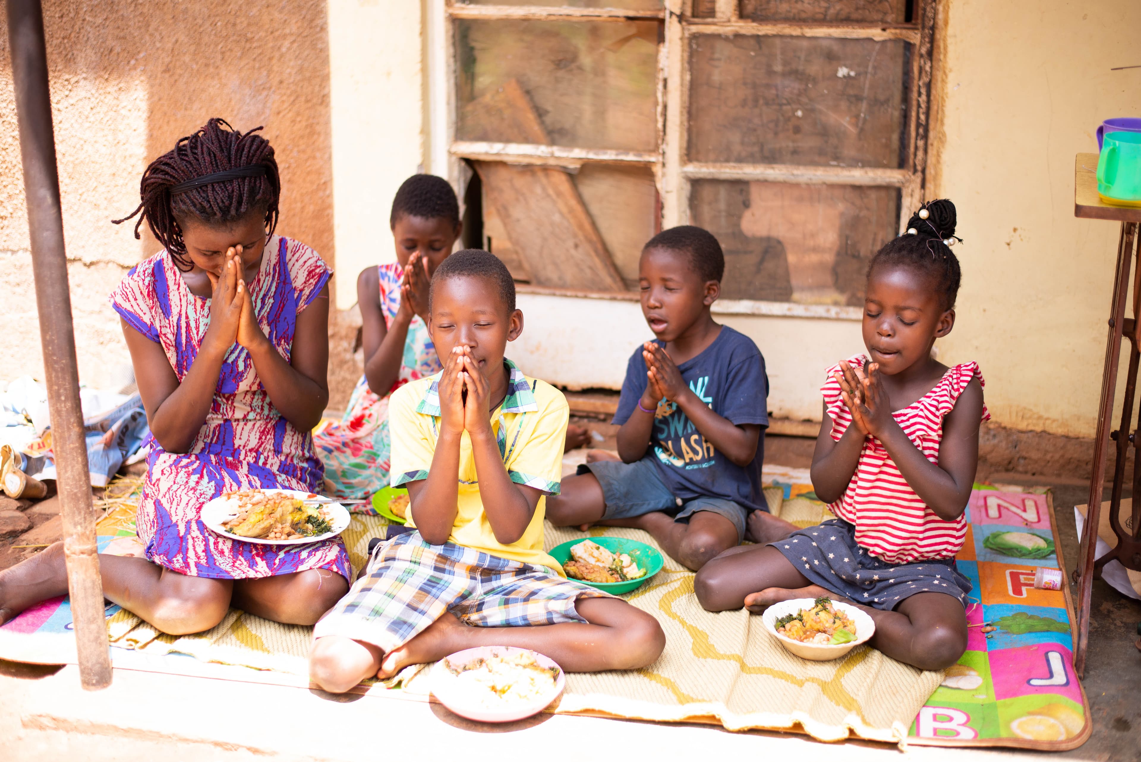 A woman wearing a purse and red dress sits on a blanket with four children. All of them have their hands folded in prayer, and there are dishes of food on the blanket with them.
