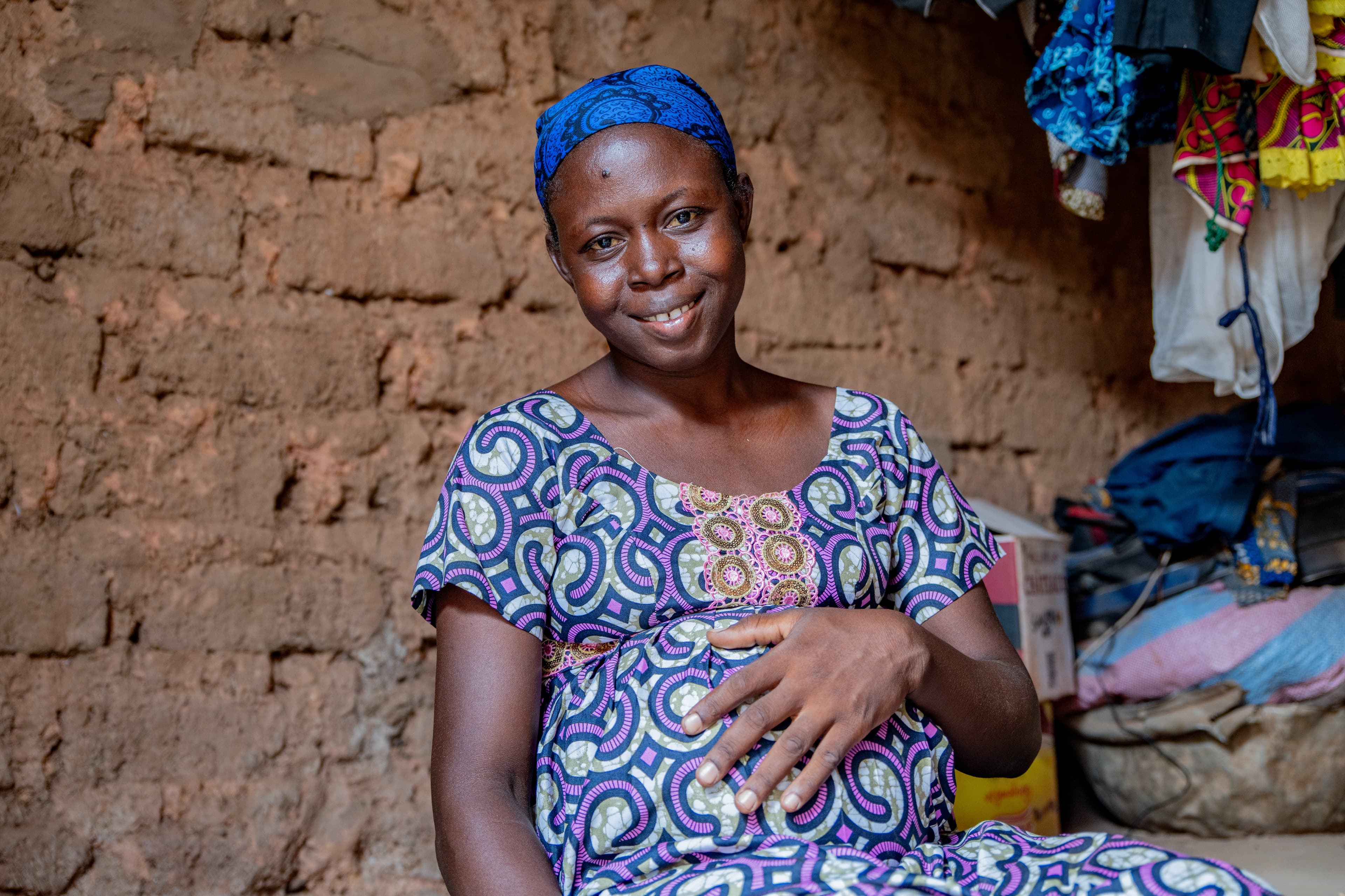 A pregnant woman sits on a mat on the floor in her sitting room holding her hand to her belly and is smiling.