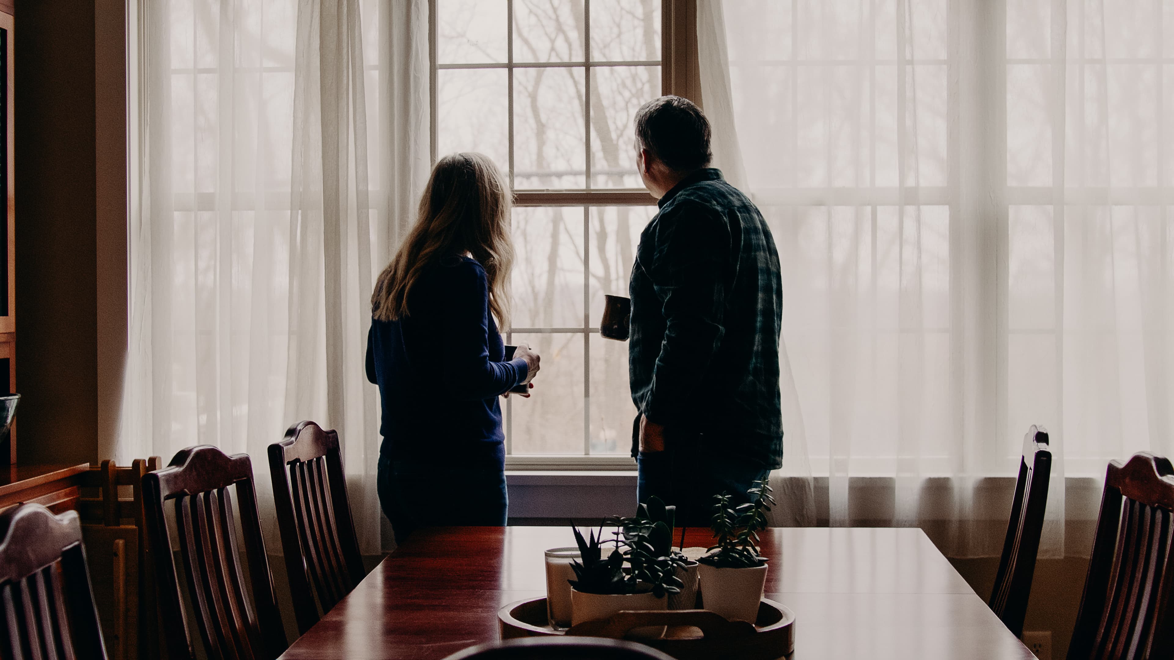 Two Compassion sponsors look out a window at their family home while holding mugs.