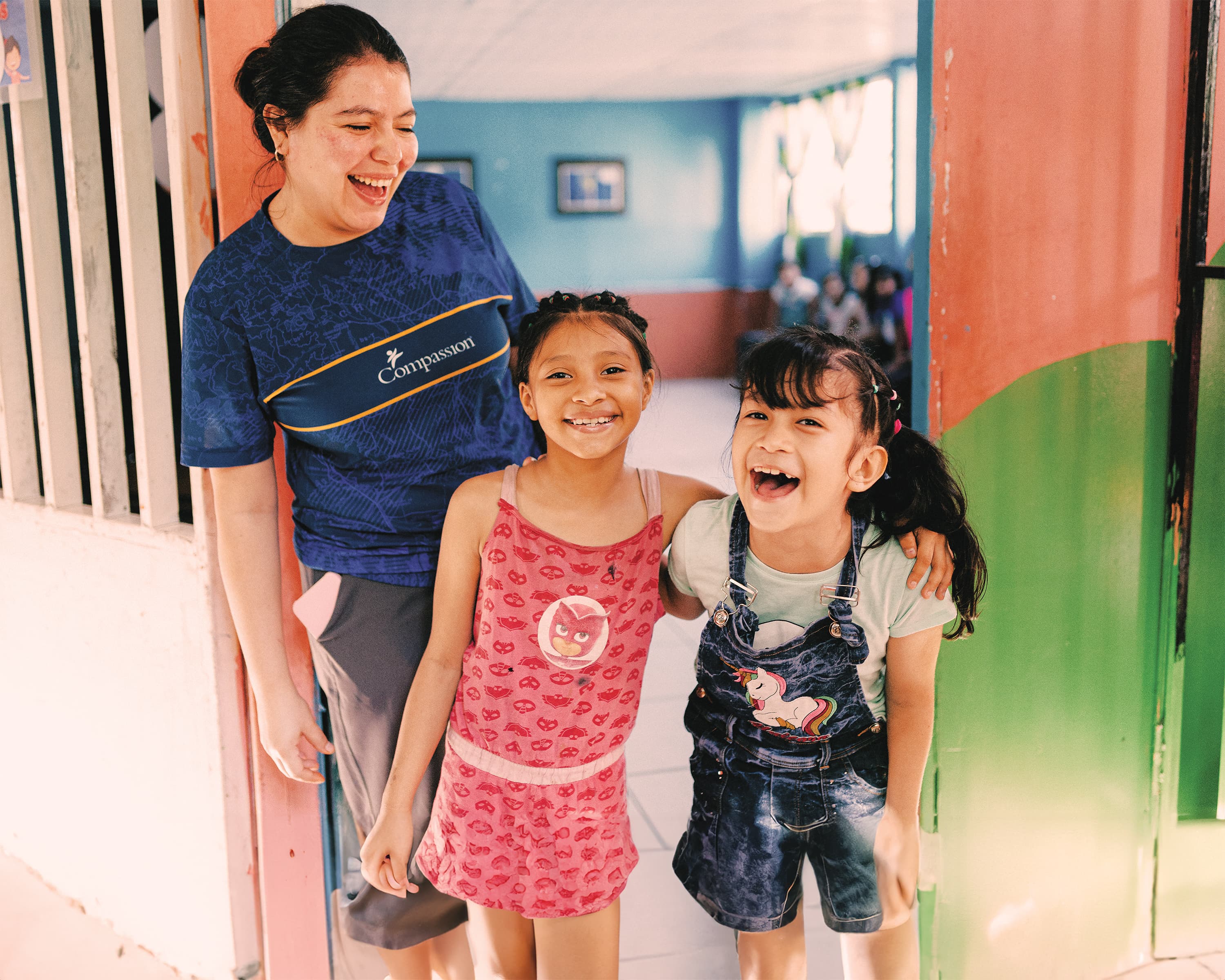 Two girls smile, stand playfully next to a woman wearing a blue Compassion shirt.