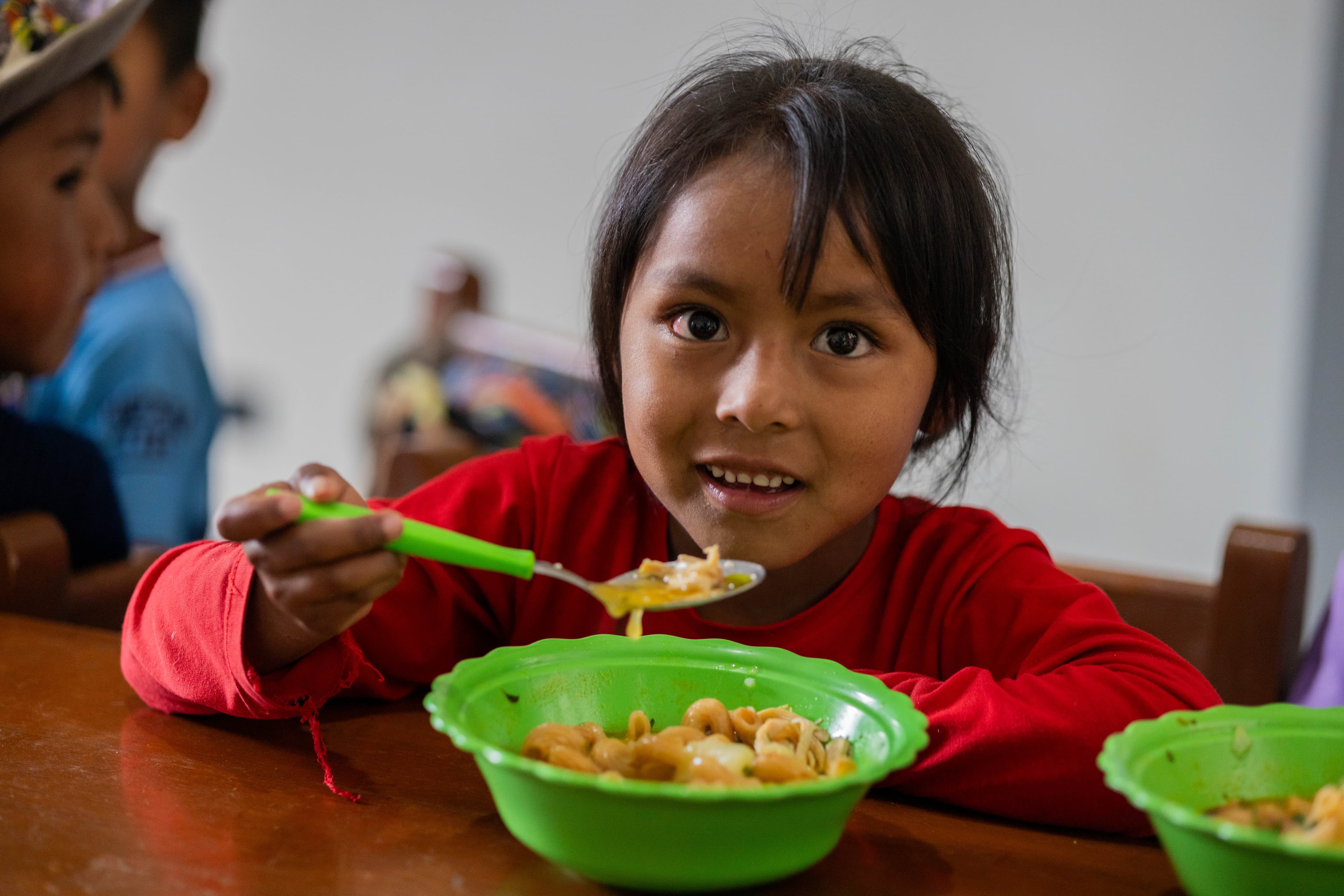 A young Bolivian girl eats from a green bowl while smiling.