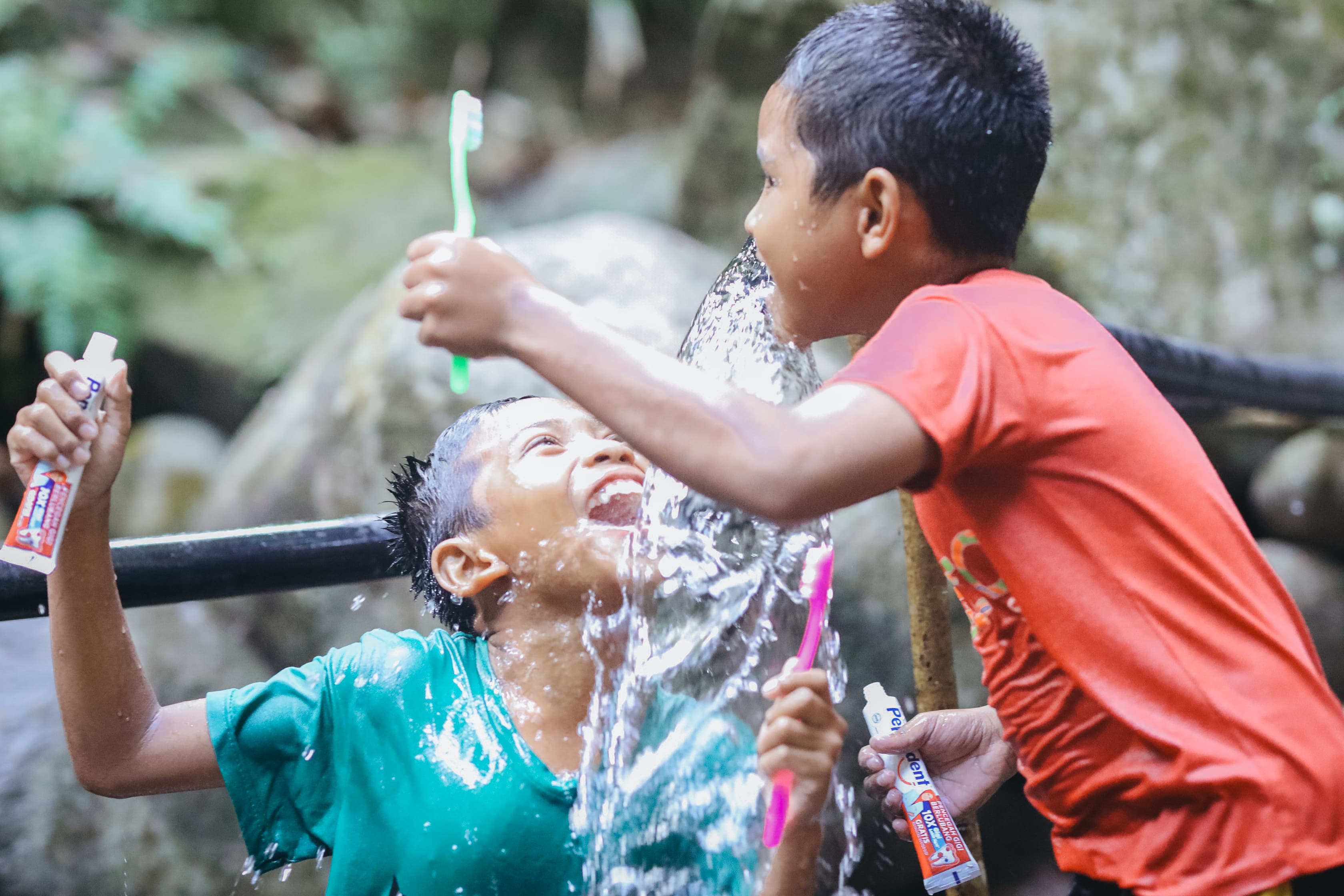 Two boys drink from a splashing water faucet while holding toothbrushes and toothpaste.