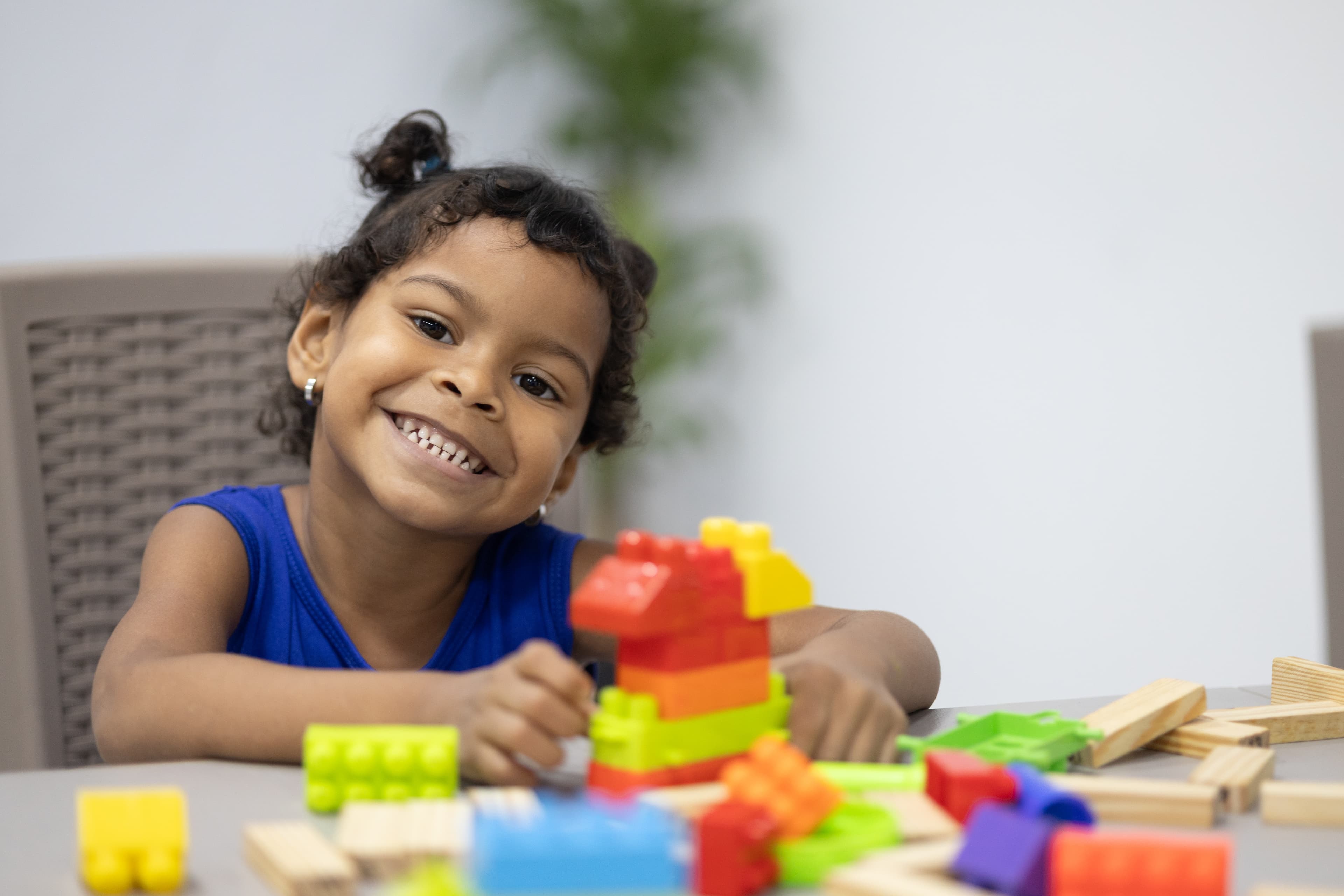 A young girl in a blue shirt sits at a tan table smiling at the camera while she works on and plays with colorful plastic interlocking blocks.