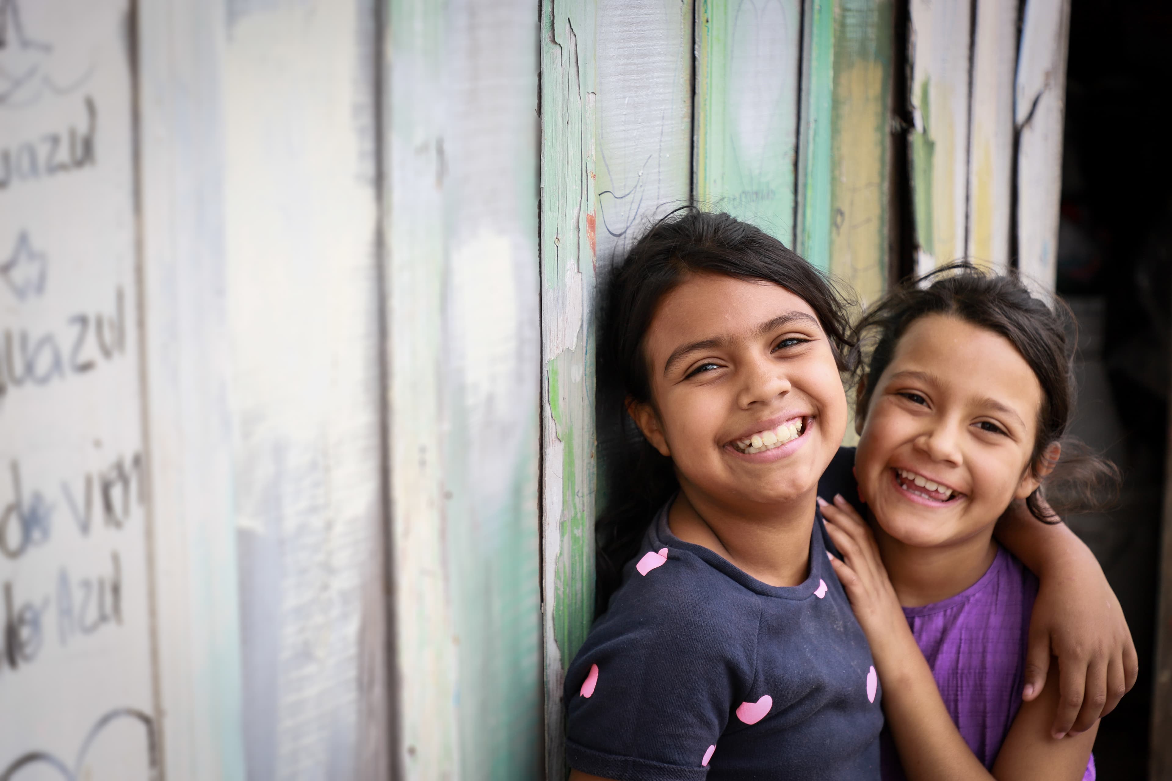 Two Honduran girls embrace and smile while leaning on a wooden wall.