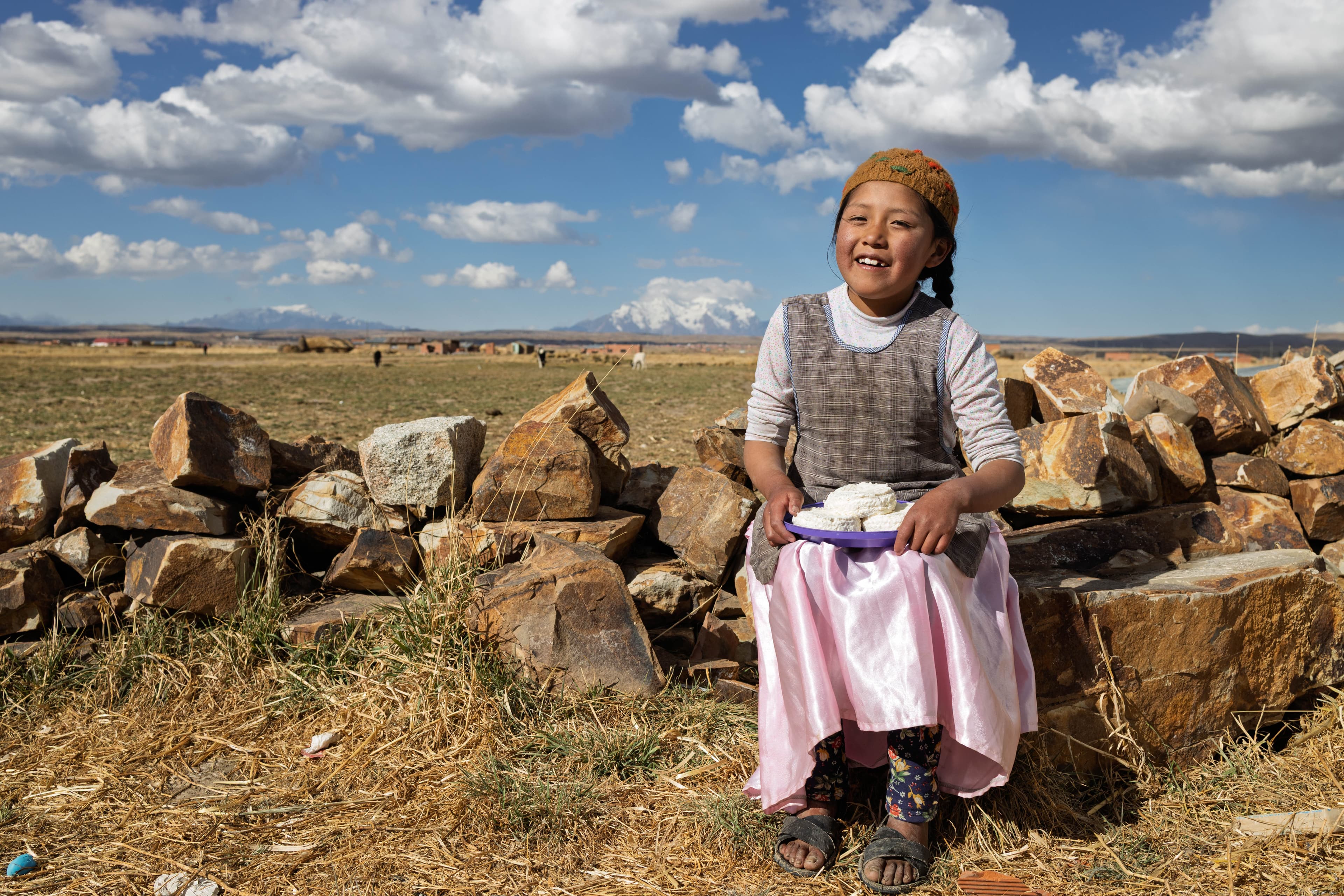 A young Bolivian girl sits on rocks while holding a plate of rice and smiling.