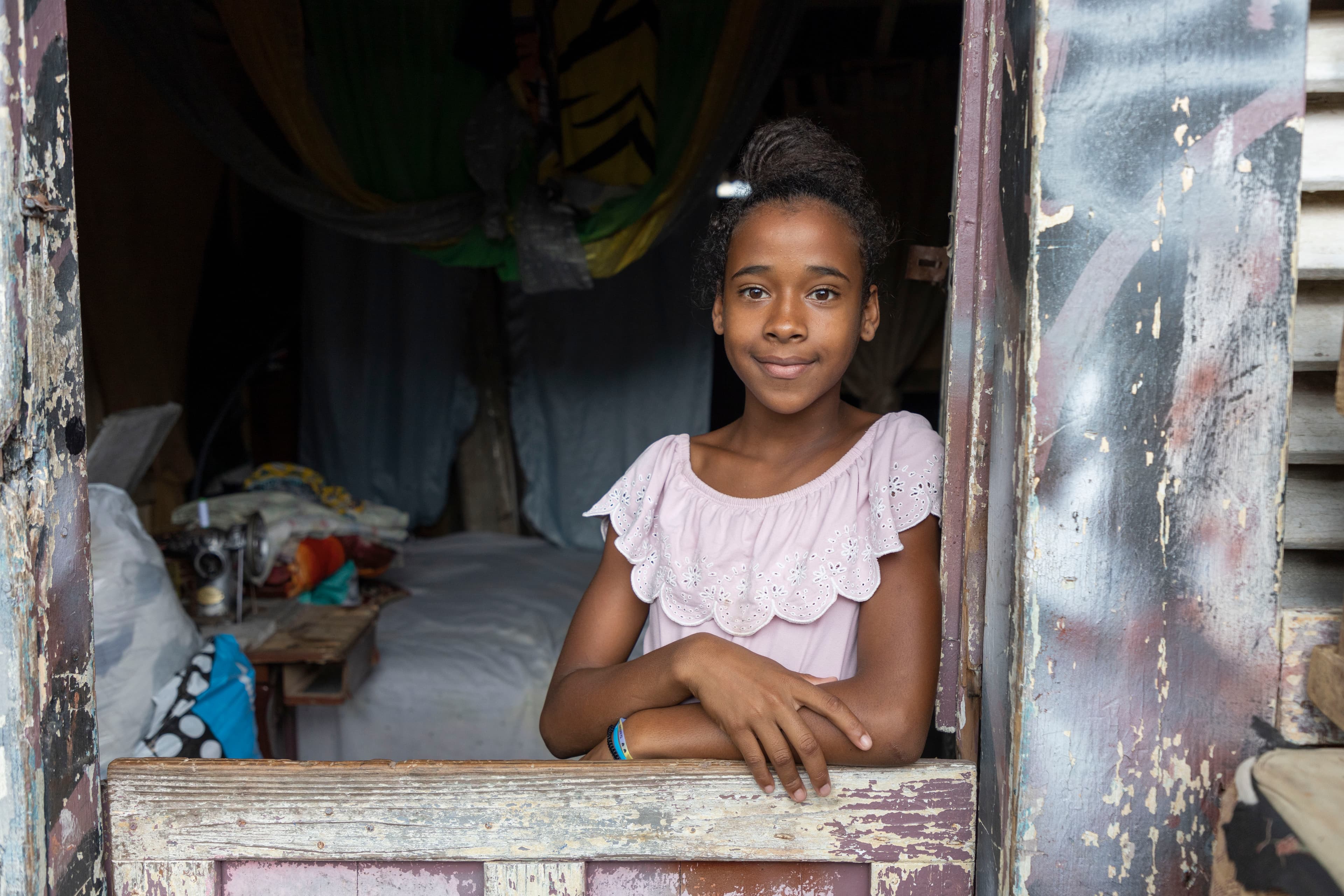 A young girl wearing a pink shirt smiles as she looks out the window of a home in the Dominican Republic.