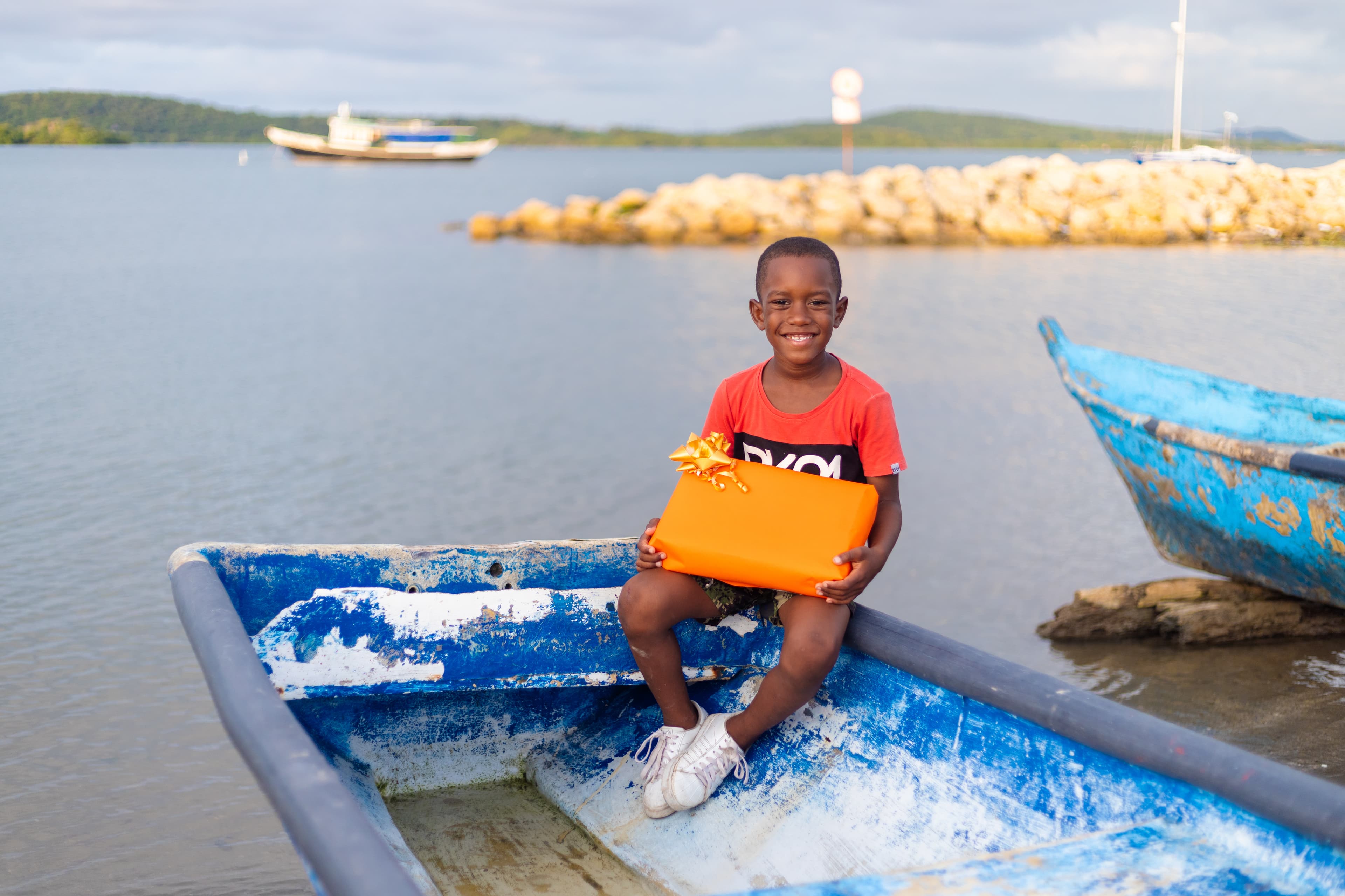 A boy sits on a boat holding a orange wrapped gift in his hands.