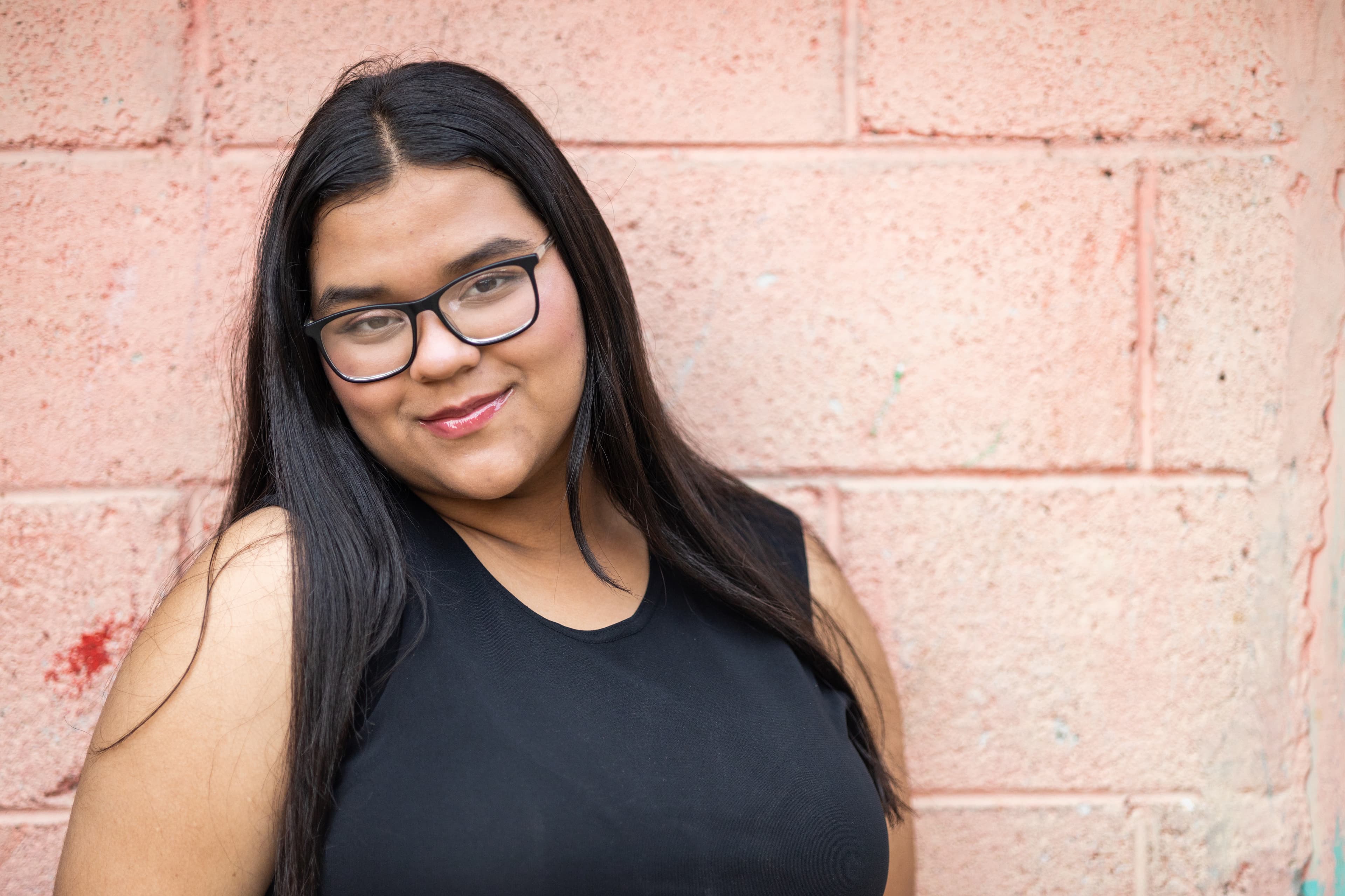 A young woman is smiling at the camera and standing in front of a pink wall.