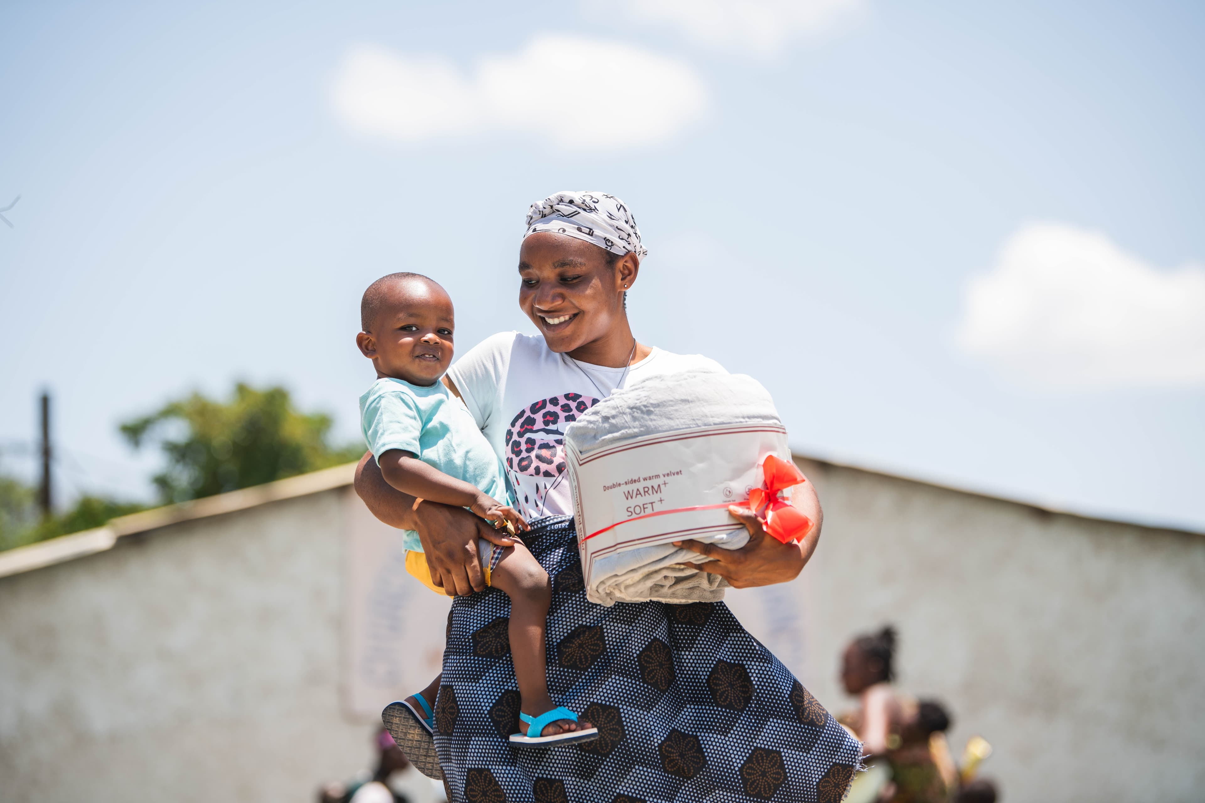 Zambian mother in a white shirt and head scarf smiles while holding her toddler son and a gifted blanket.