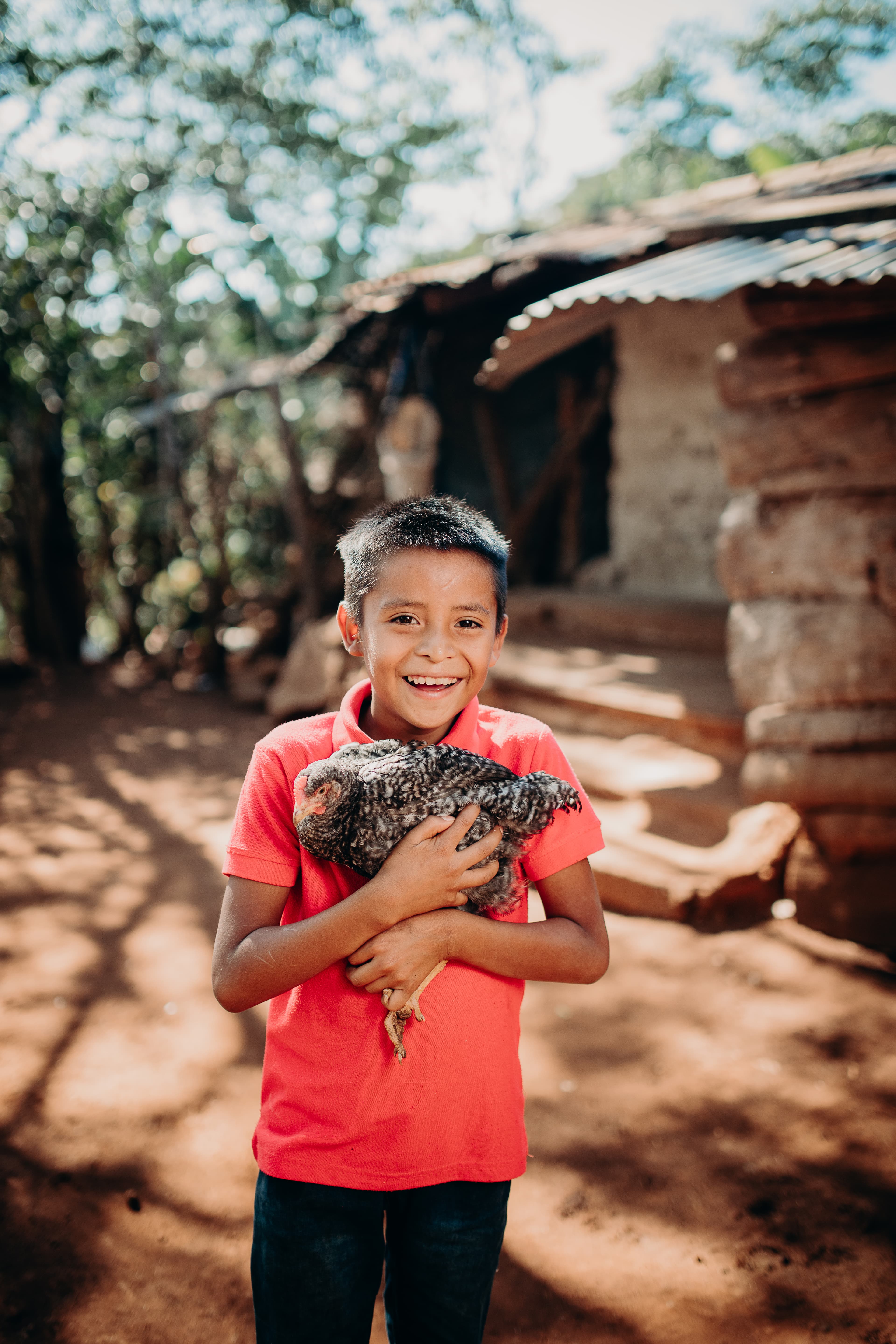 Boy smiles while holding his family’s chicken in front of his home.