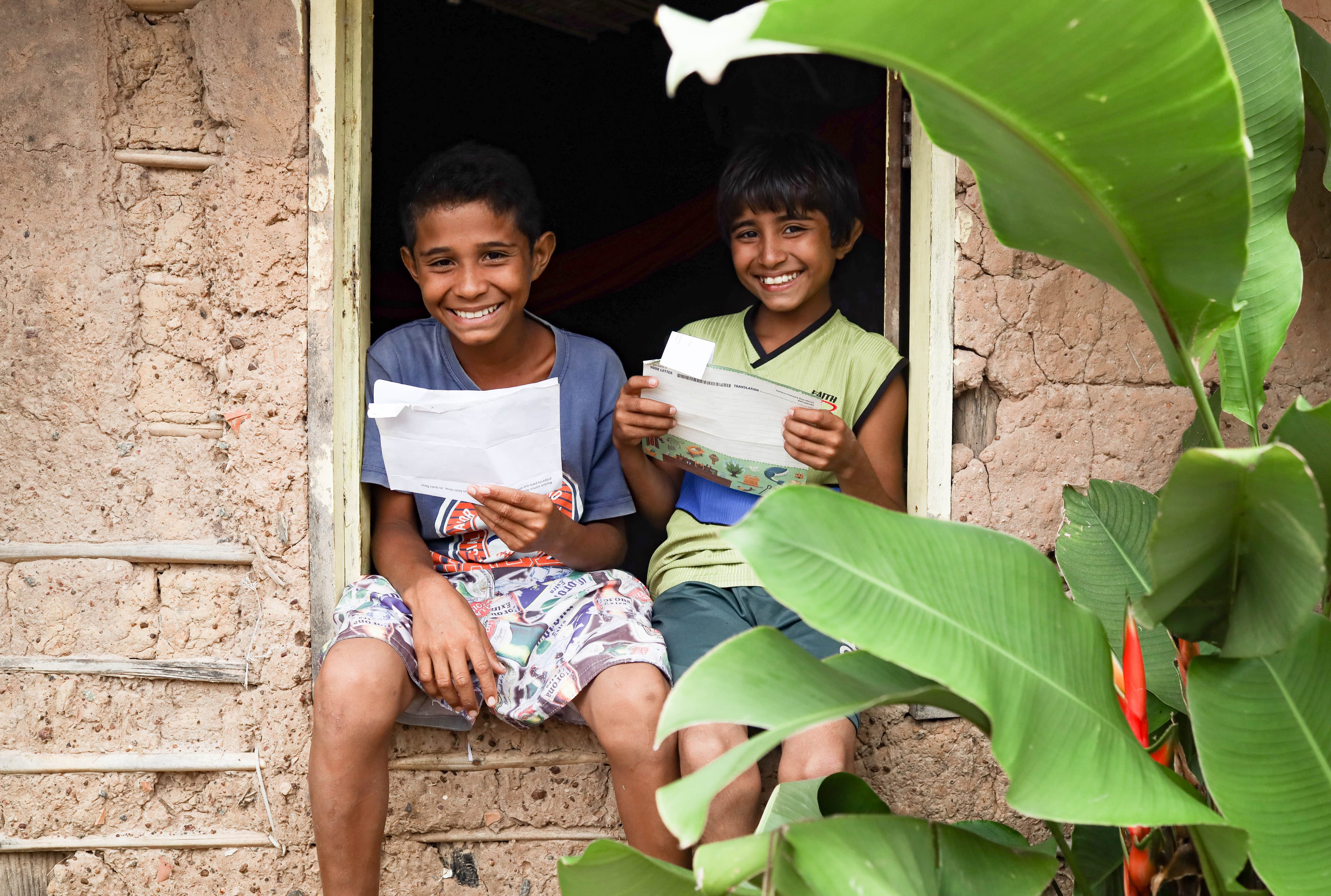 Two smiling, young boys hold sponsorship letters while sitting on a window sill