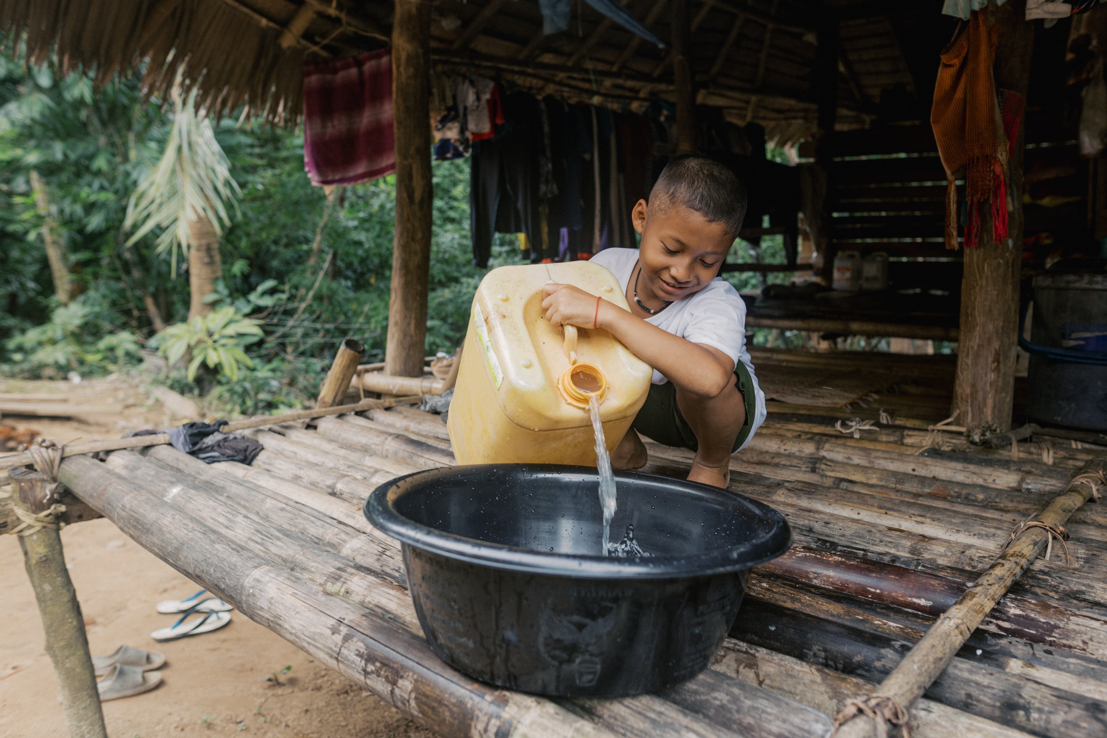 Young, Thai boy squats on a bamboo deck while pouring water from a large jug into a black basin.