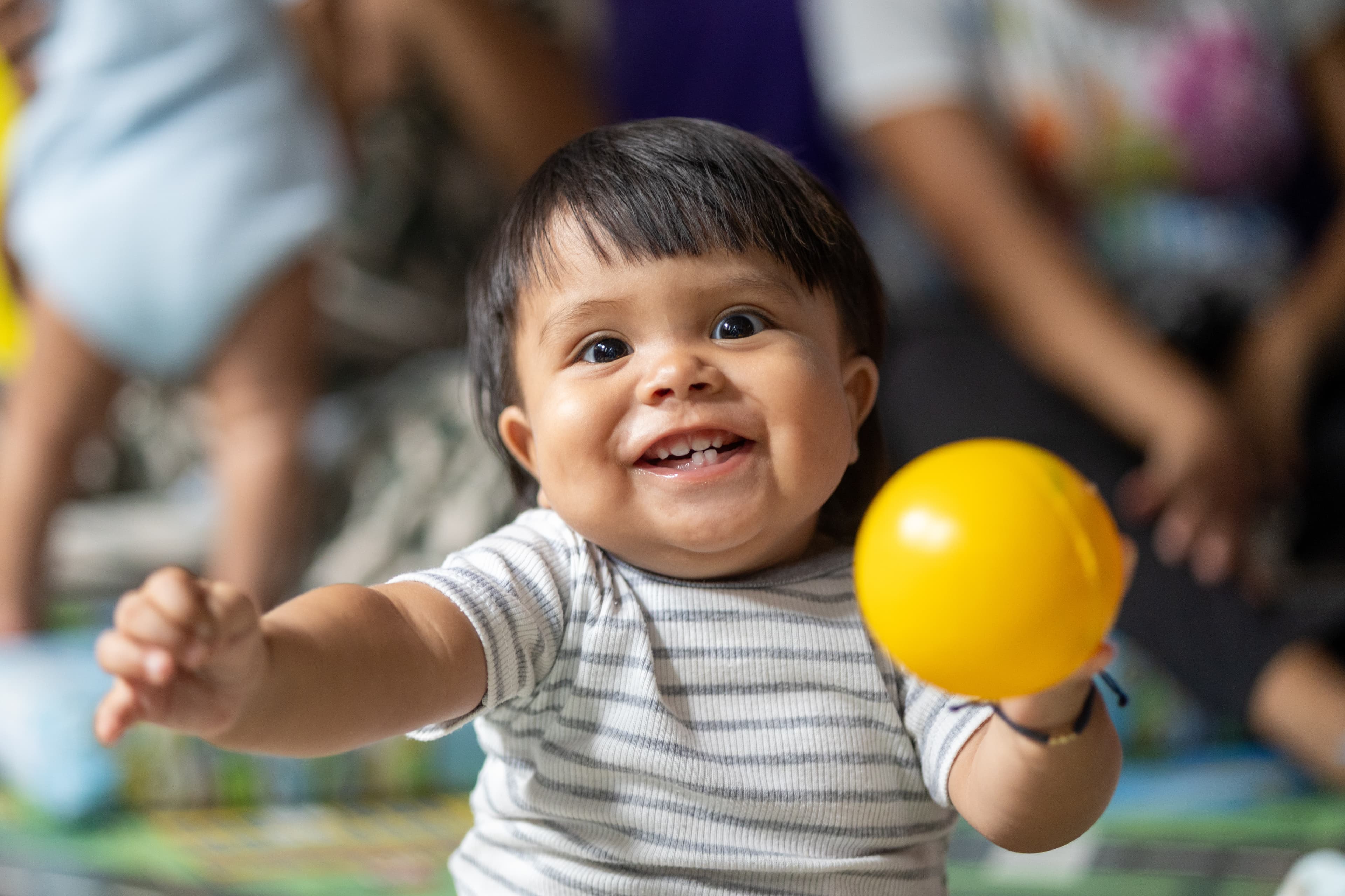 A baby boy is smiling, holding a yellow ball in one hand and reaching out with the other.