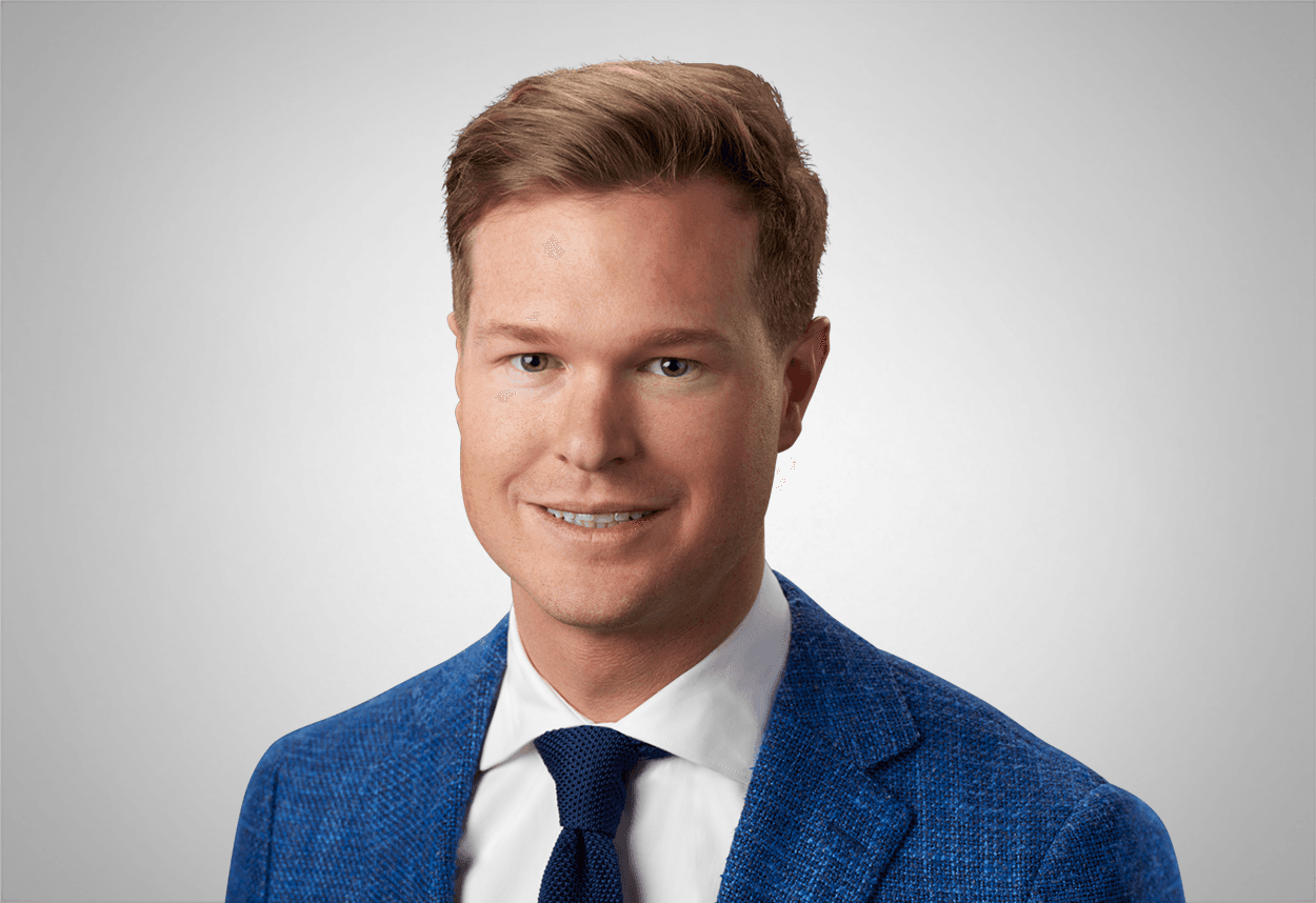 A man in a blue tweed suit jacket, white shirt and navy blue tie is smiling, with a grey background behind him.