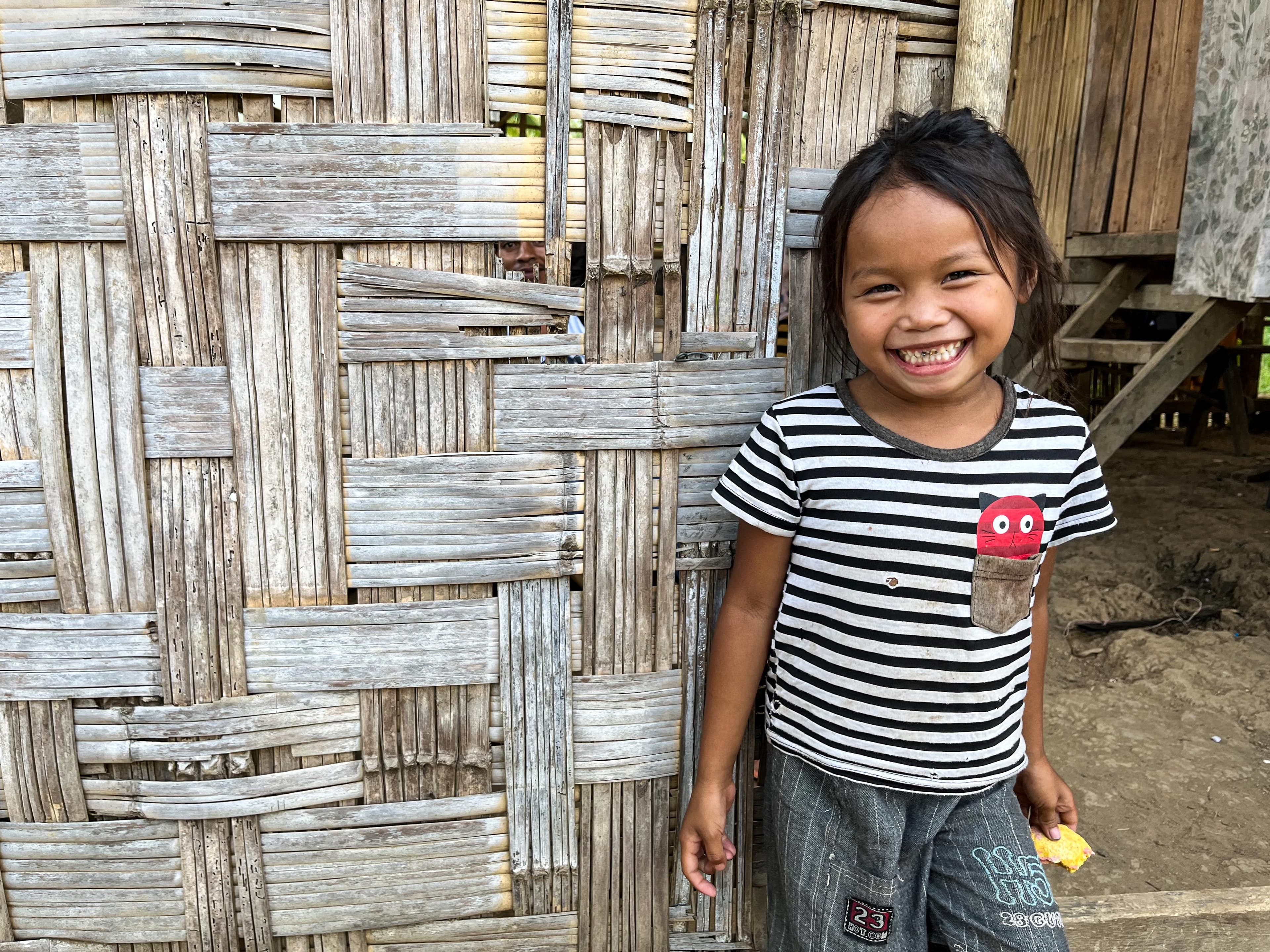 A girl in the Philippines is wearing a striped shirt and smiling outdoors in front of a bamboo wall.