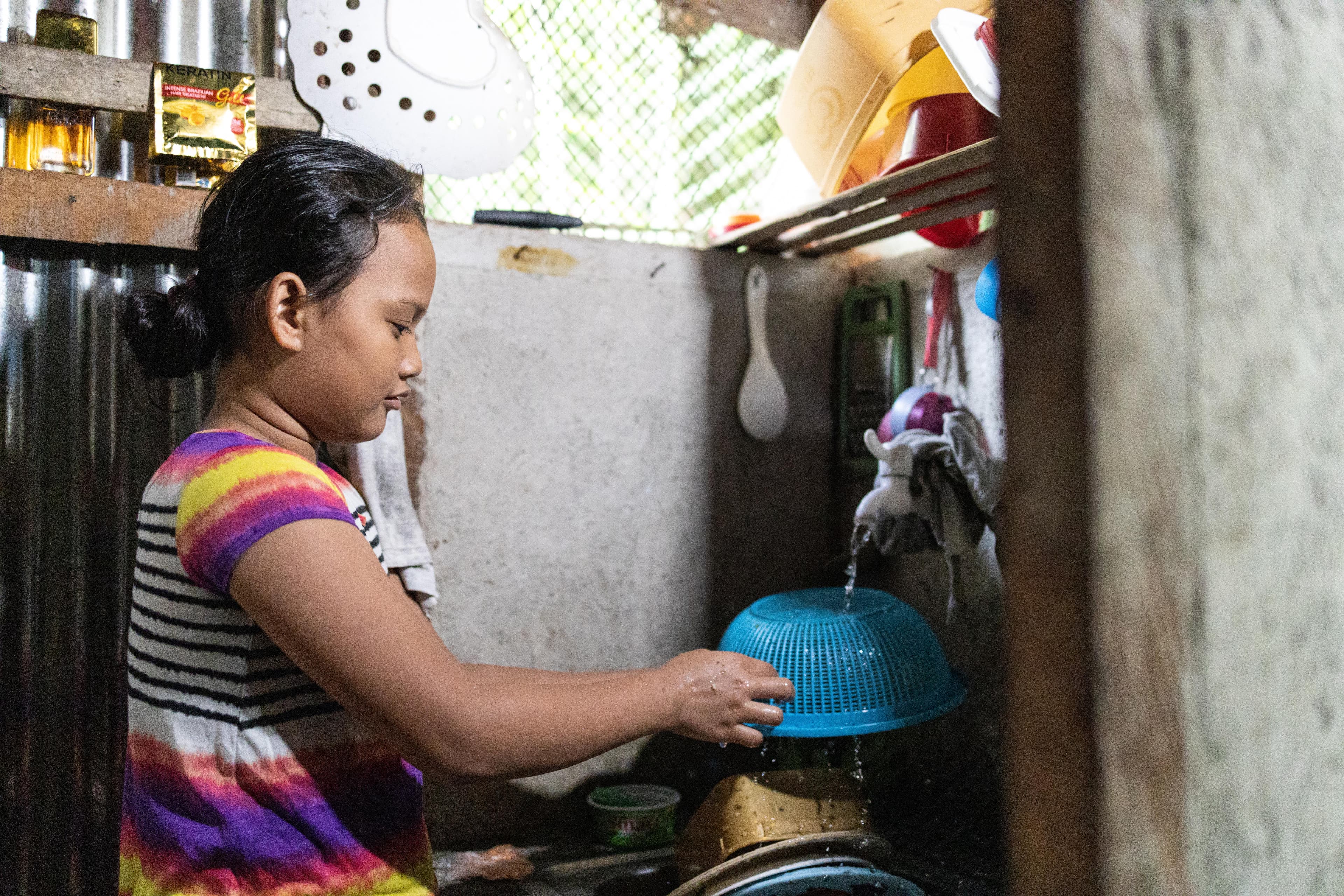 A young girl washes a blue dish in a tiny sink.