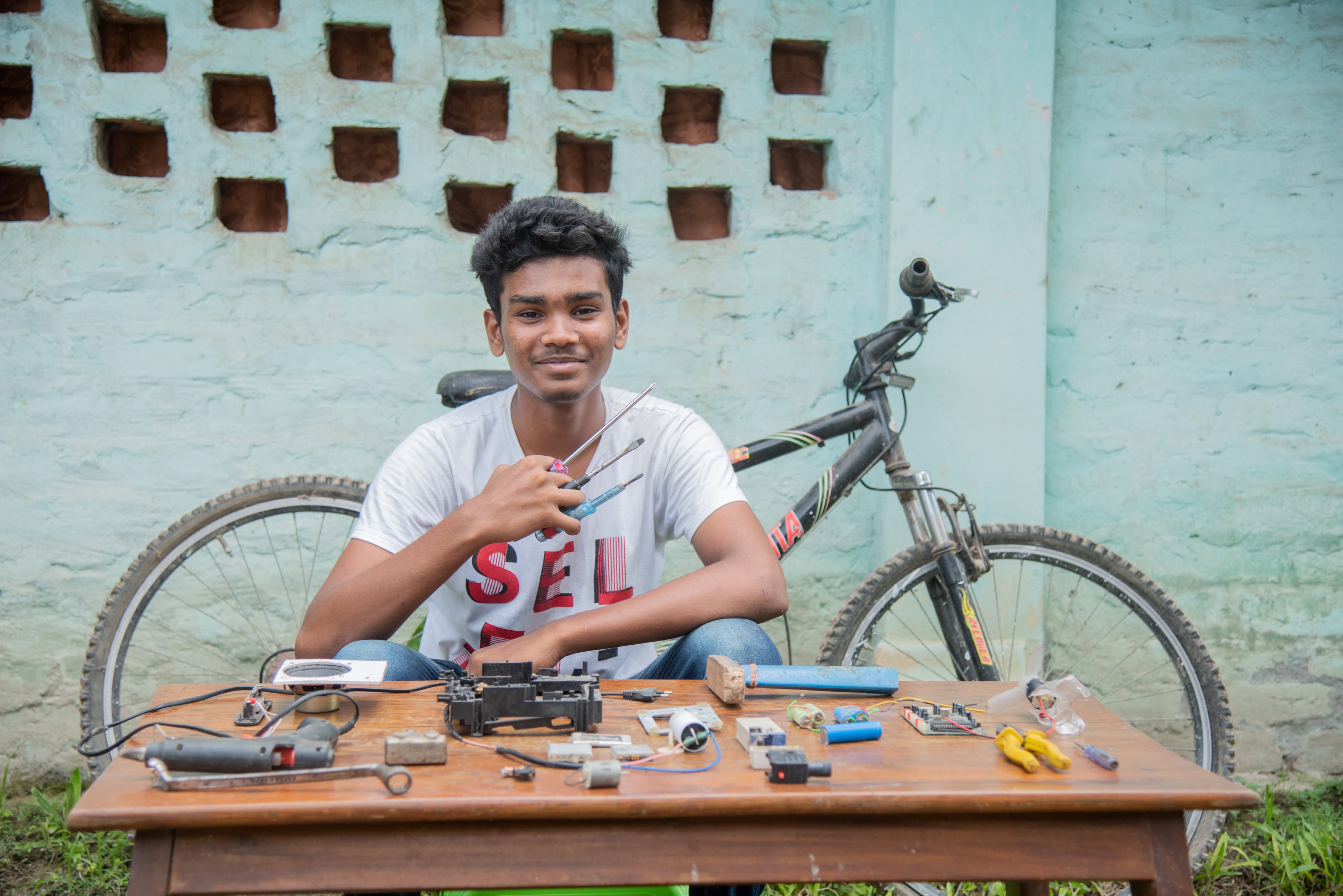 Teenaged boy sits in front of a bicycle with a table and tools.