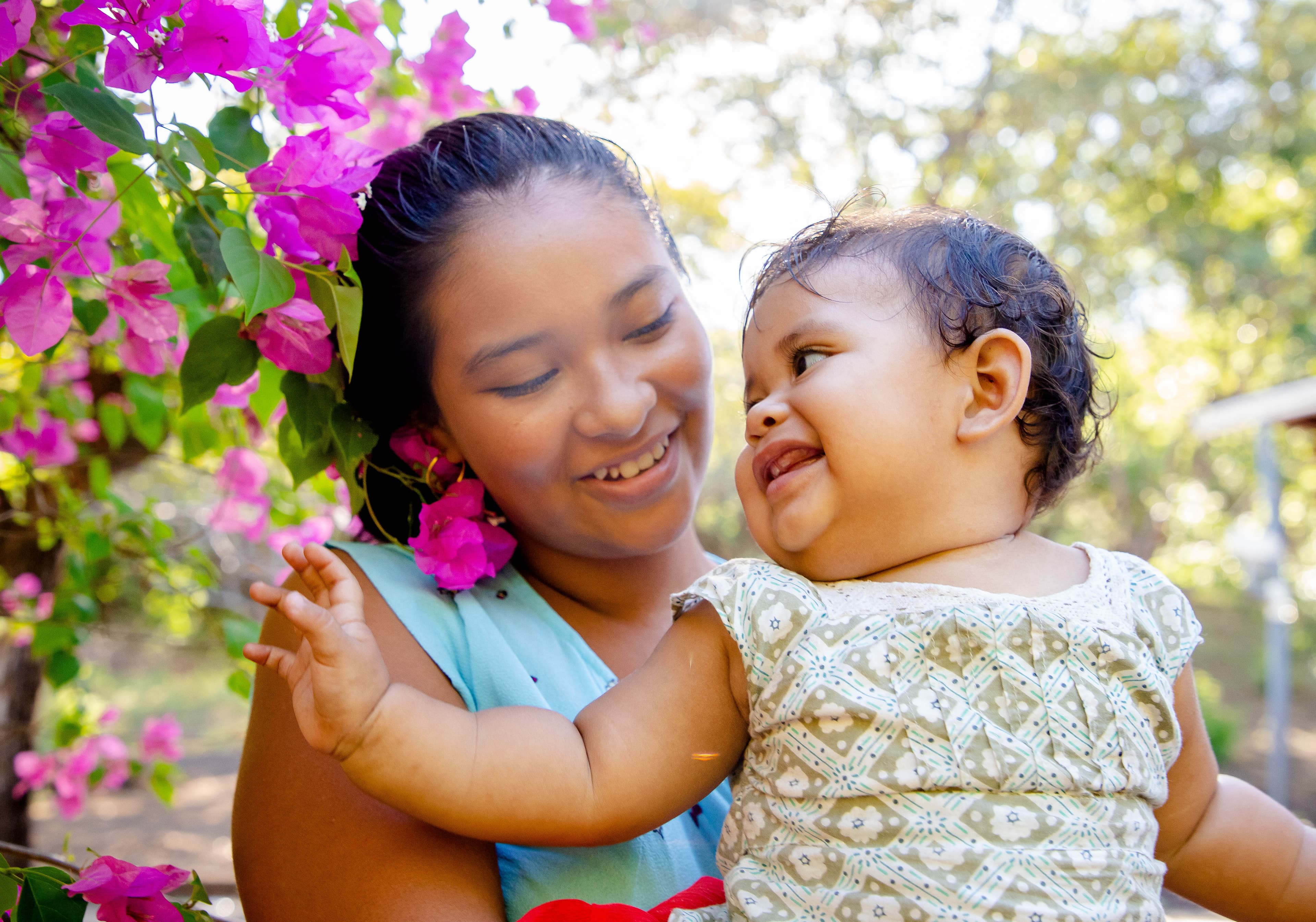A mother is holding her baby and smiling as the baby reaches for a flower.