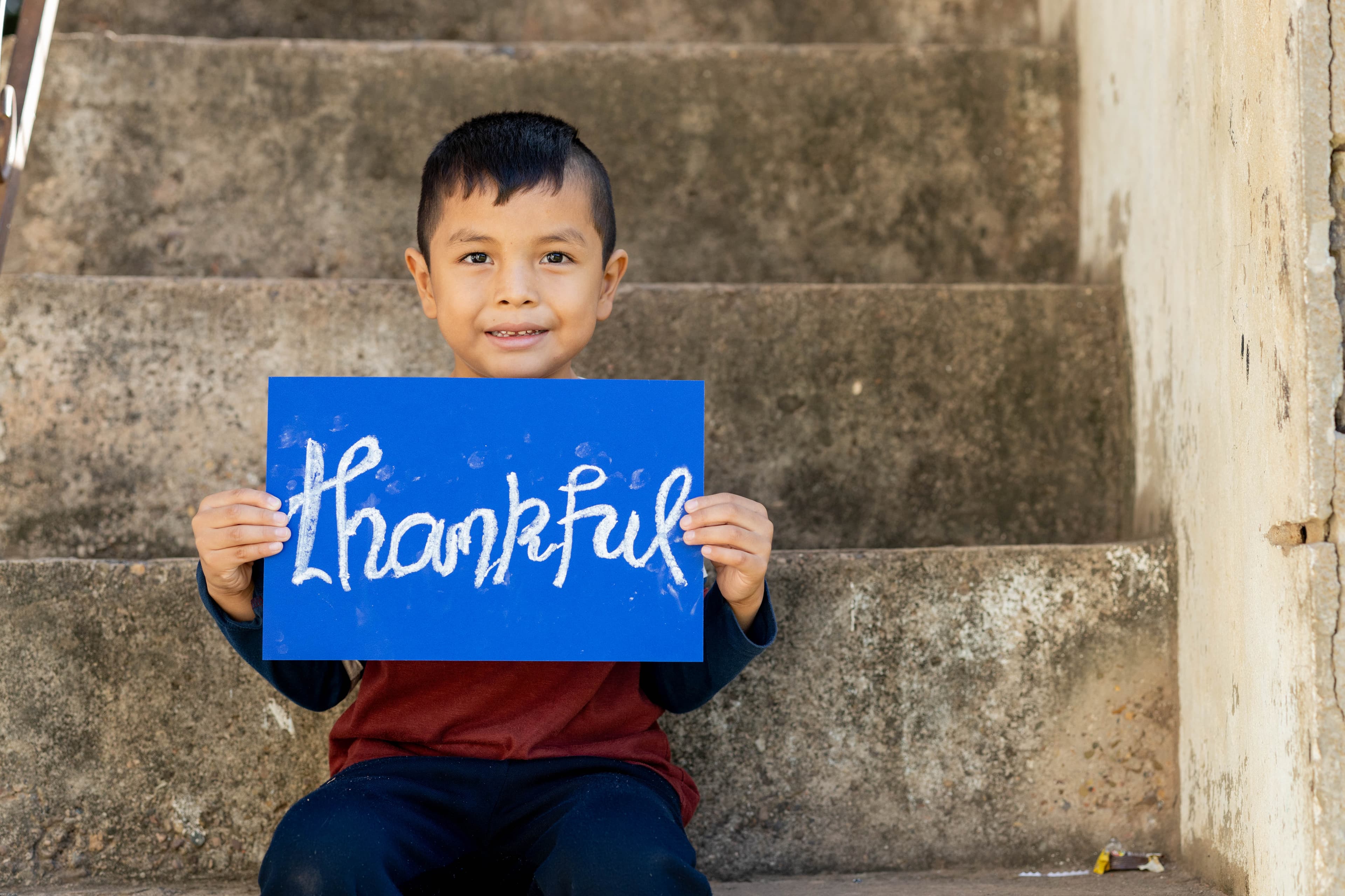A young boy sits on concrete steps while holding a sign that says thankful.