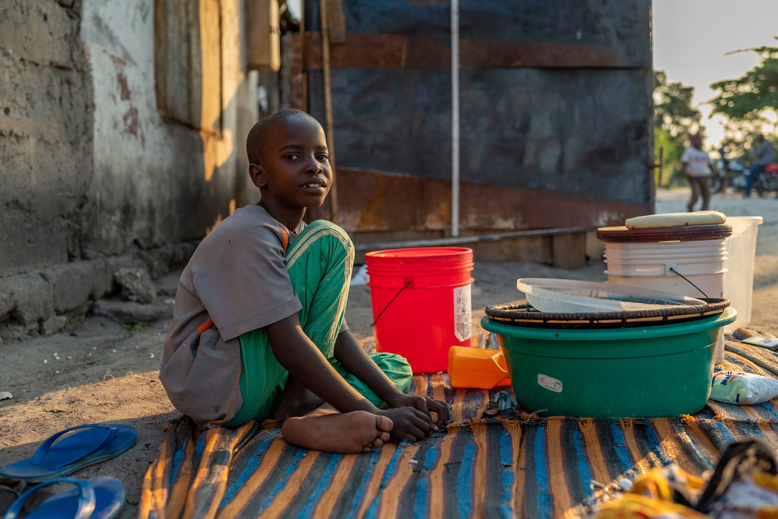 A teenage boy sits barefoot on a blanket outdoors. Buckets and other supplies are beside him.